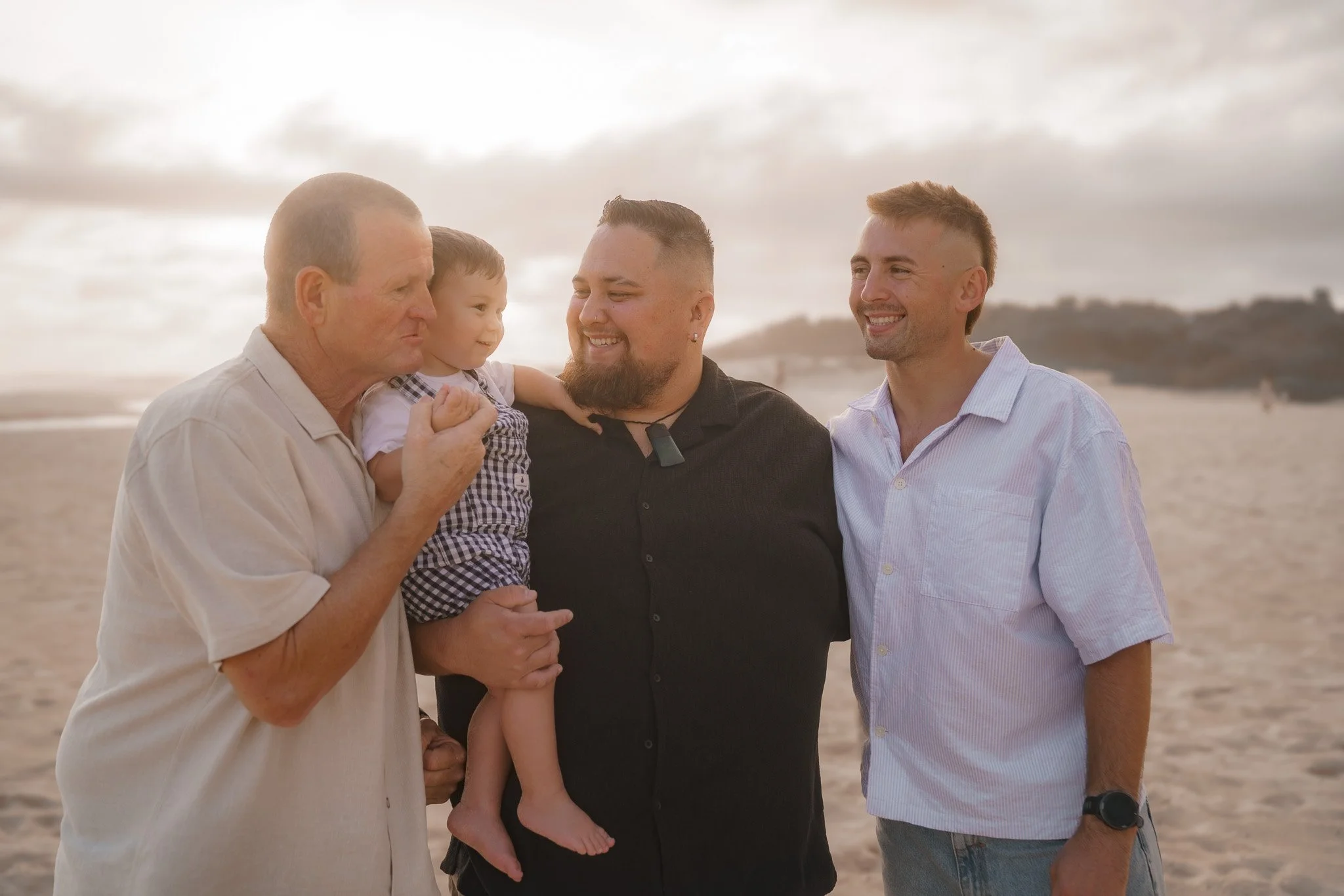 Four men, including a young child, standing on the beach during sunset, smiling and looking at each other.