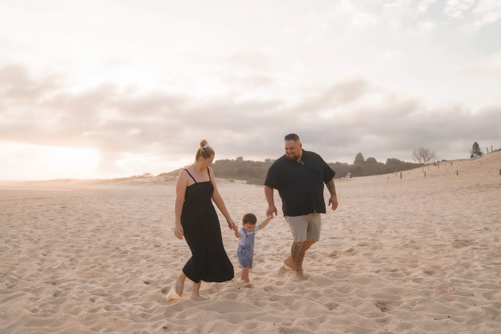 A family of three enjoying a walk on the beach during sunset, holding hands. The woman is wearing a black dress, the man is in a black shirt and beige shorts, and the child is in a blue and white checkered outfit.