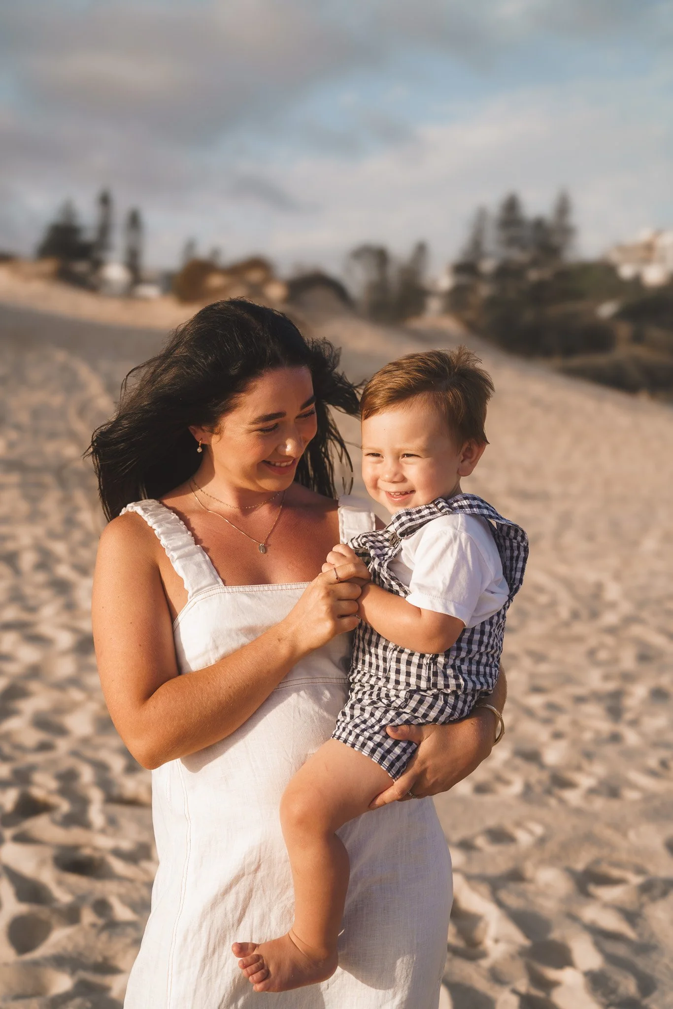 A woman with long dark hair smiling and holding a young boy with short light brown hair, wearing shorts and a checkered shirt, on a sandy beach during daylight.