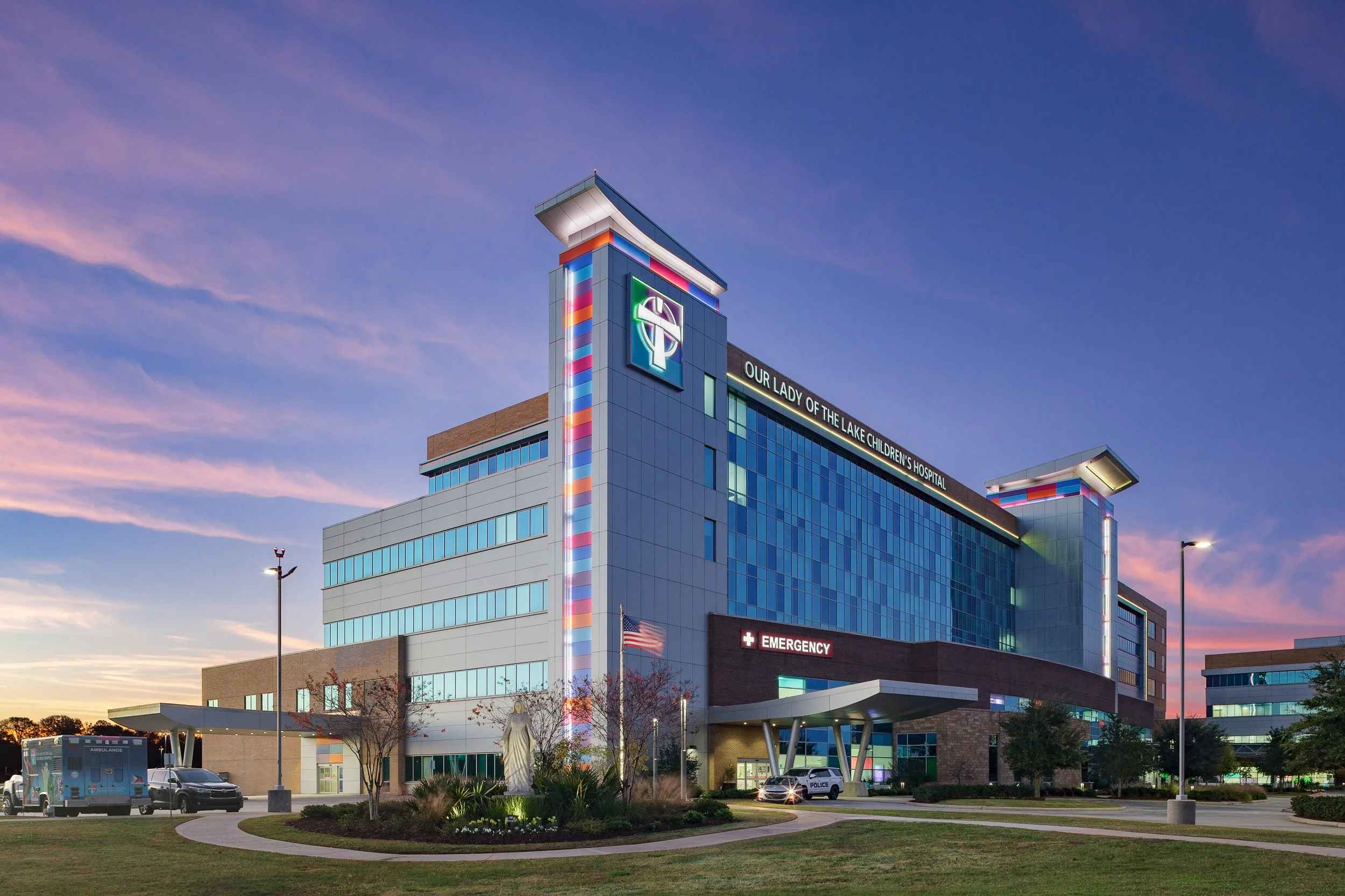 Modern hospital building with colorful lighting, ambulance and police vehicles outside, at dusk.