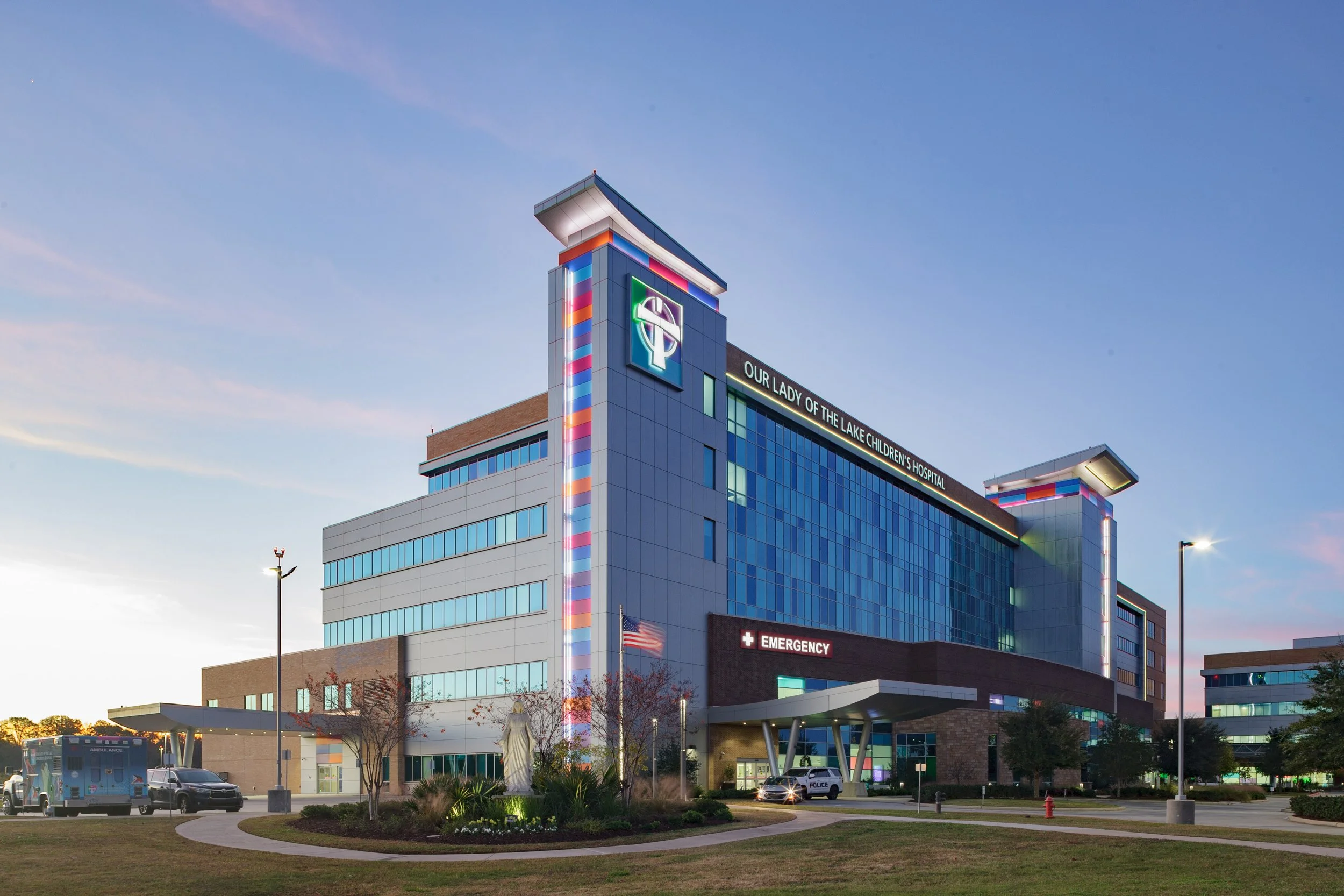 Exterior view of Our Lady of the Lake Children's Hospital, a modern multi-story building with blue glass windows, colorful vertical light strips, and a sign indicating emergency services. The hospital's name and a logo are visible at the top. The sur
