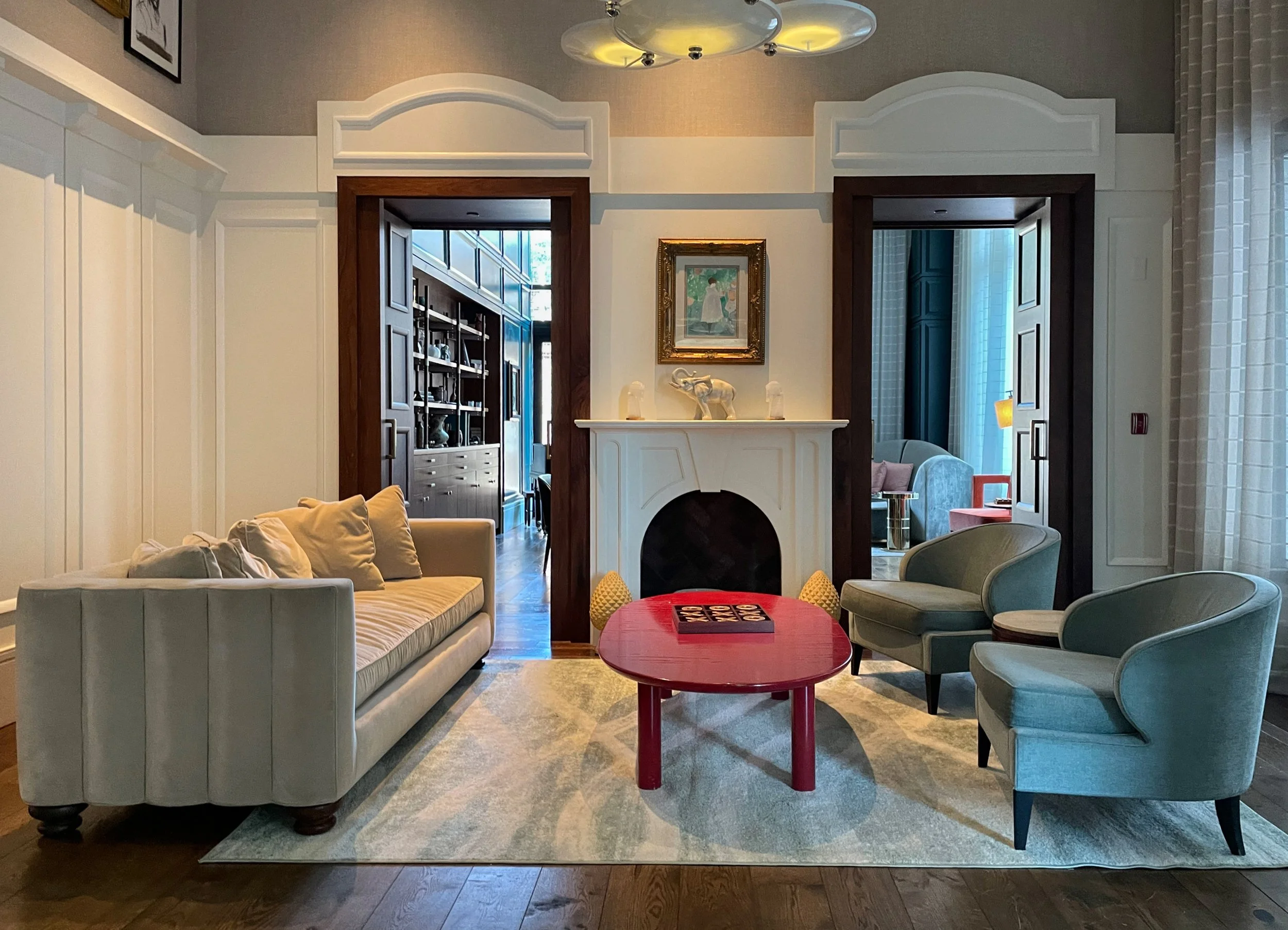 Living room with beige sofa, two light green armchairs, a red coffee table, a white fireplace with decorative sculptures, and artwork above it. Two doorways lead to other rooms, with built-in shelving visible through the left doorway. The room has a