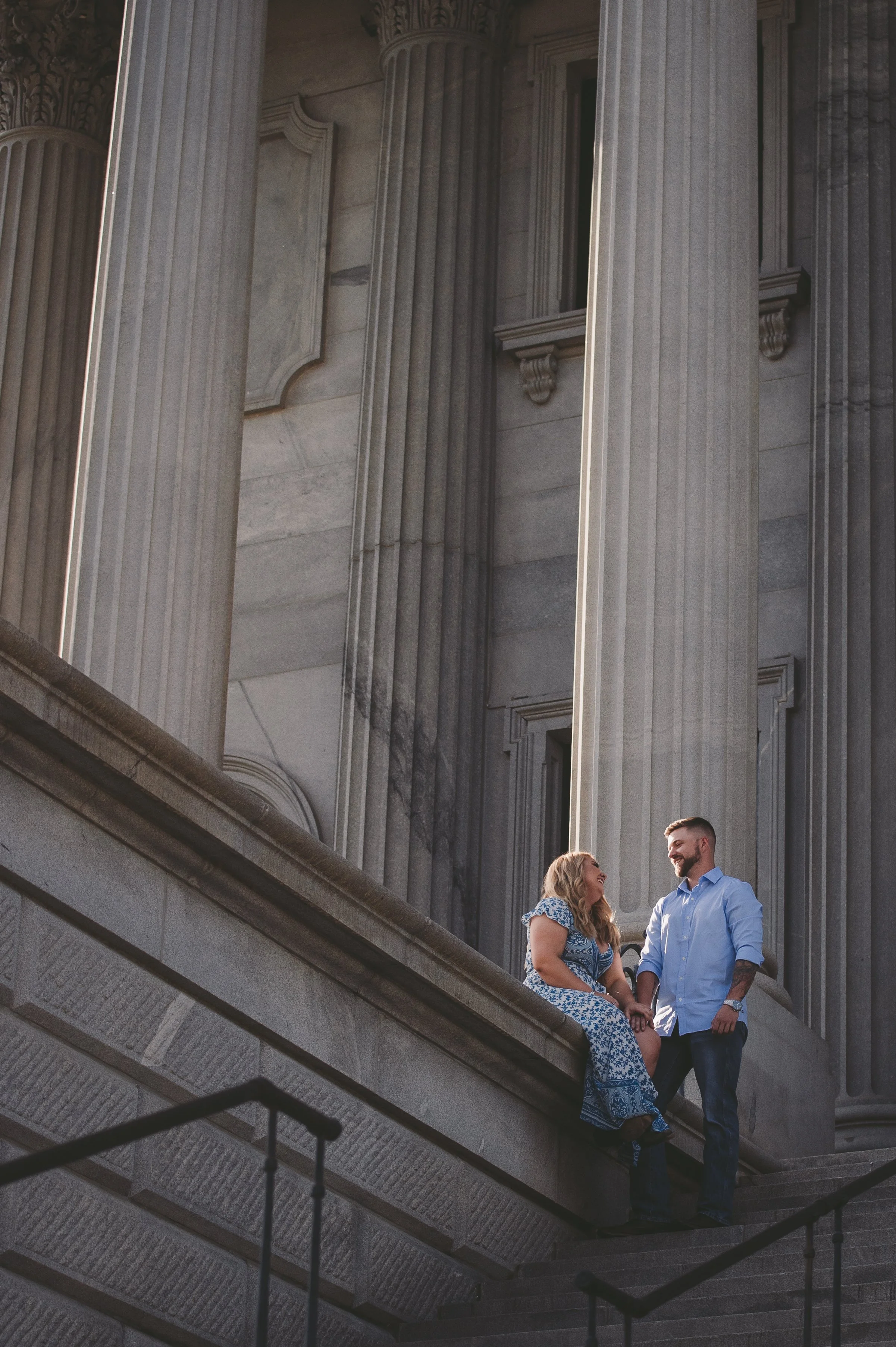 A woman and man standing and sitting on steps outside a building with large classical columns, engaging in conversation and smiling.