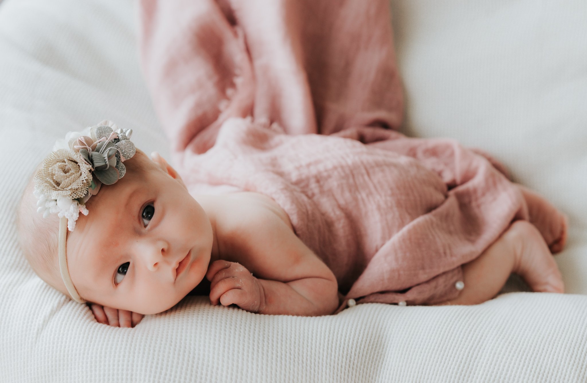 A baby girl lying on a white textured blanket, wearing a floral headband and wrapped in a pink swaddle.