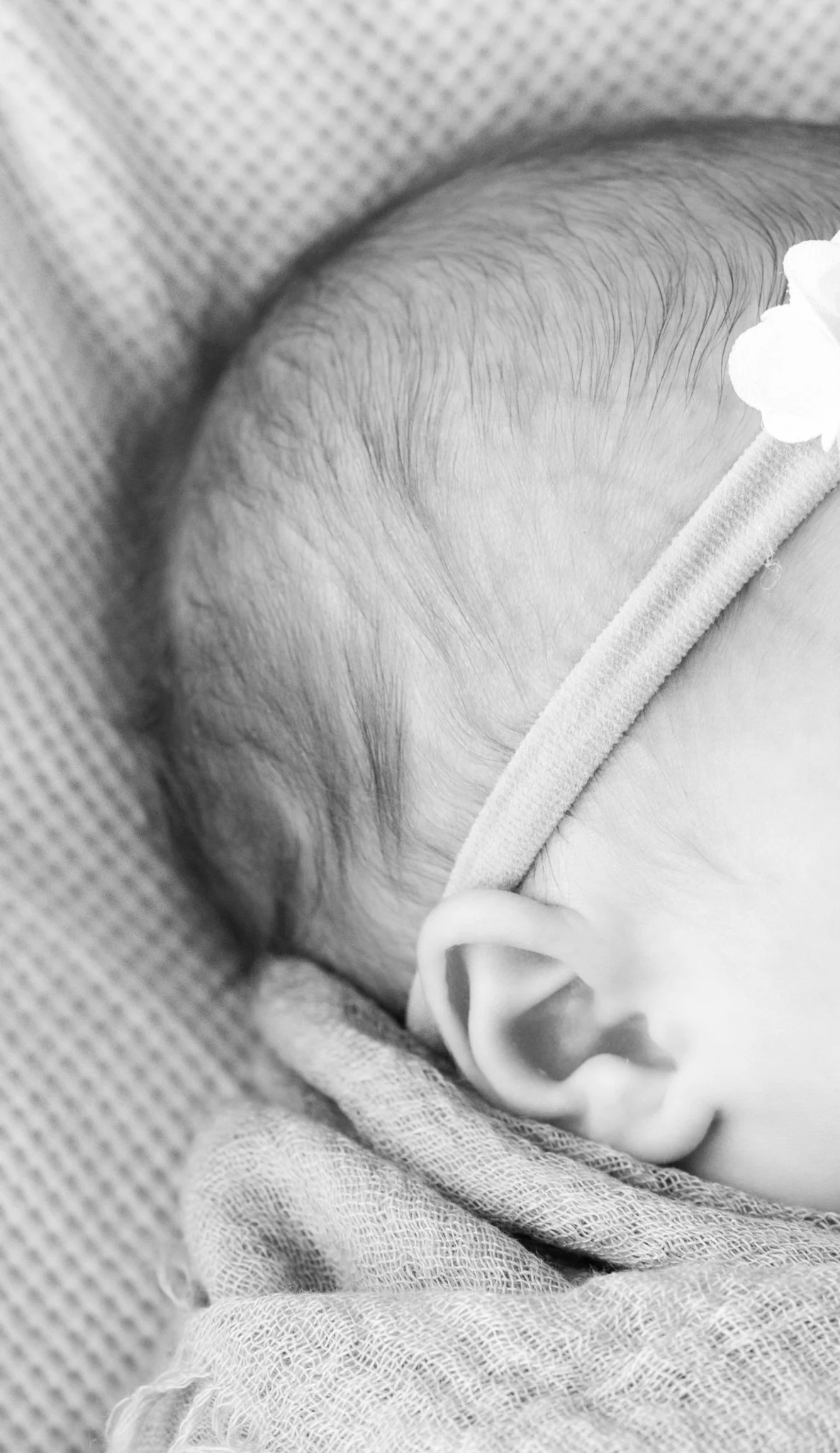 Close-up of a sleeping baby's head, showing fine hair and ear, wrapped in a blanket with a floral headband.