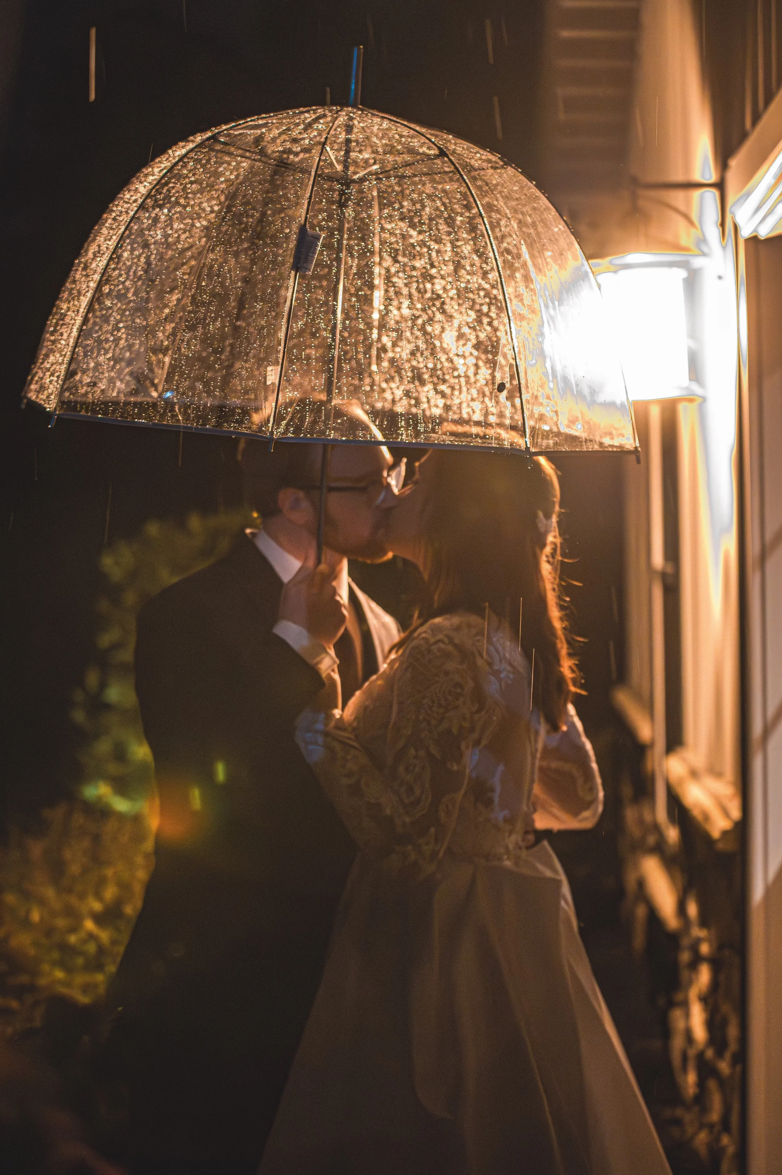 A couple dressed in formal attire kissing under a clear umbrella with rain droplets at night, illuminated by a nearby light.