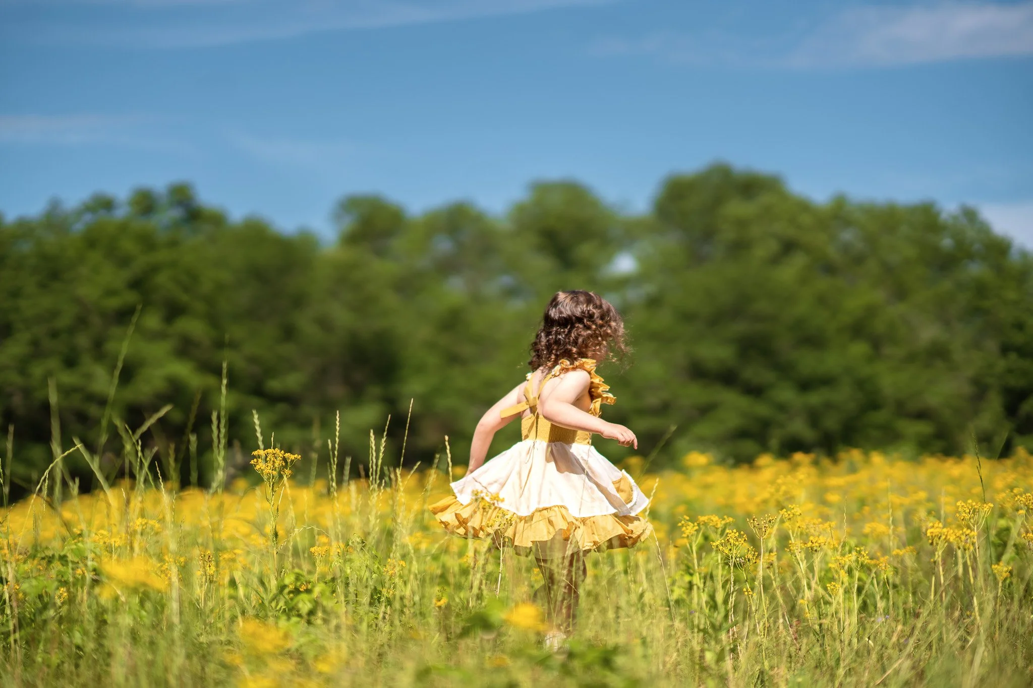 A young girl with curly brown hair in a yellow dress with ruffled sleeves and a layered skirt running through a field of yellow flowers, with green trees and a blue sky in the background.