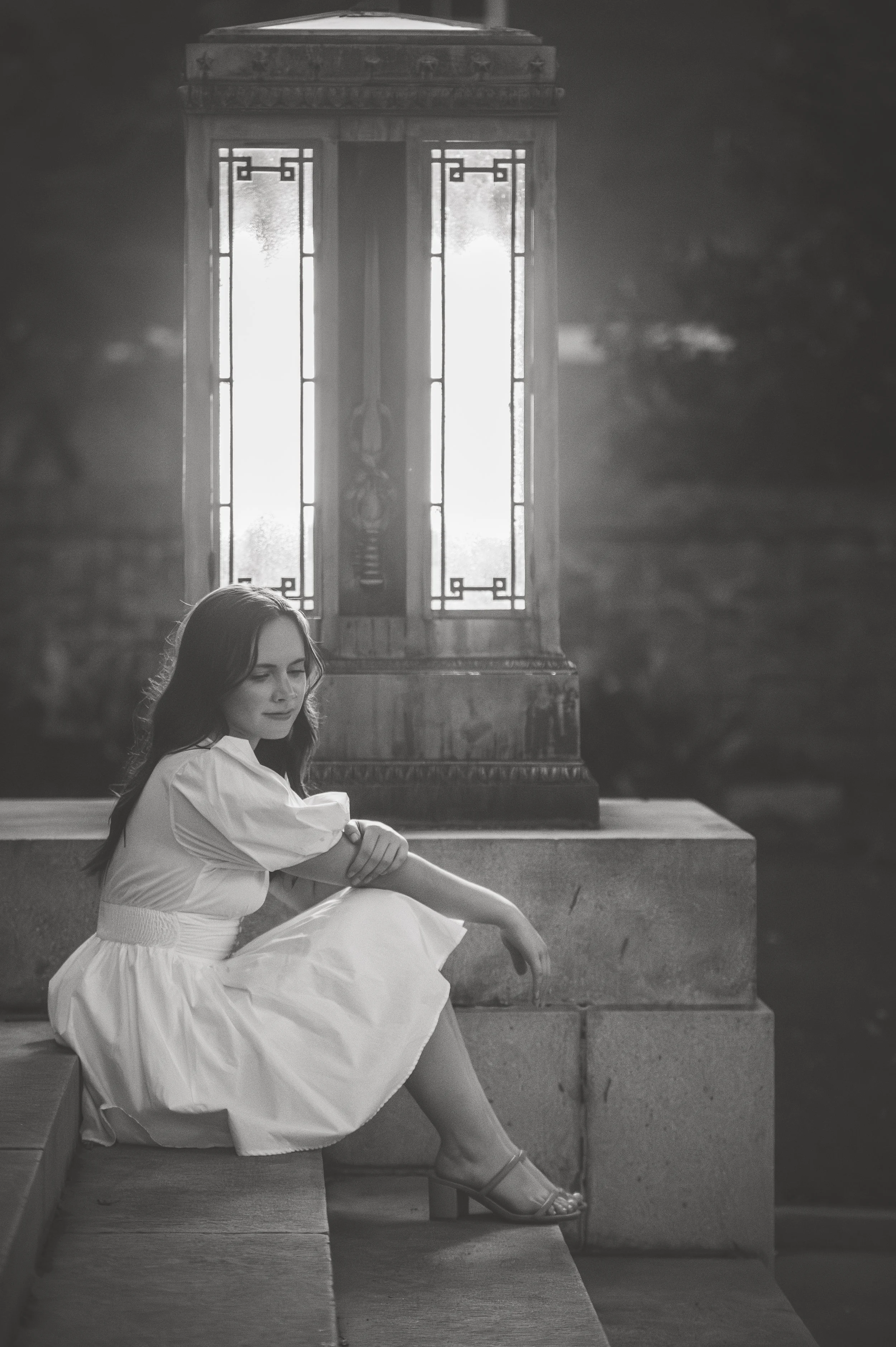A young girl in a white dress sitting on steps near a wooden structure with stained glass panels, in black and white.