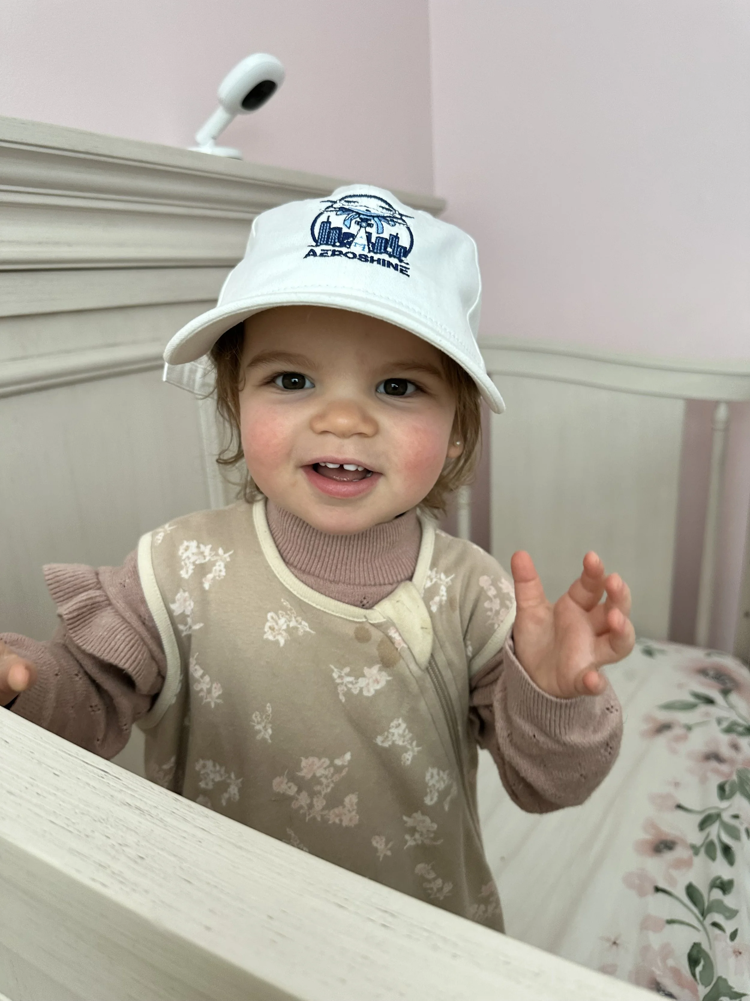 A young girl smiling and looking at the camera, wearing a white bucket hat with blue embroidery and a beige floral jacket, standing by a white bed with floral bedding.