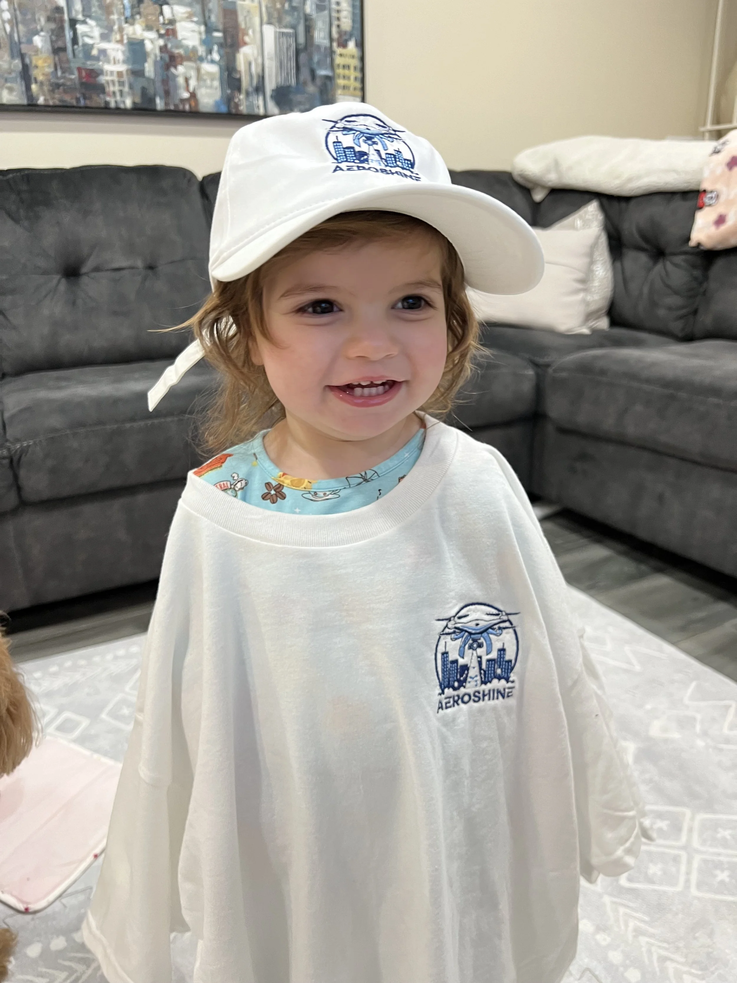 A young child with curly hair wearing a white Aeroshine cap and a white Aeroshine T-shirt, smiling in a living room with a dark sofa and a patterned rug.