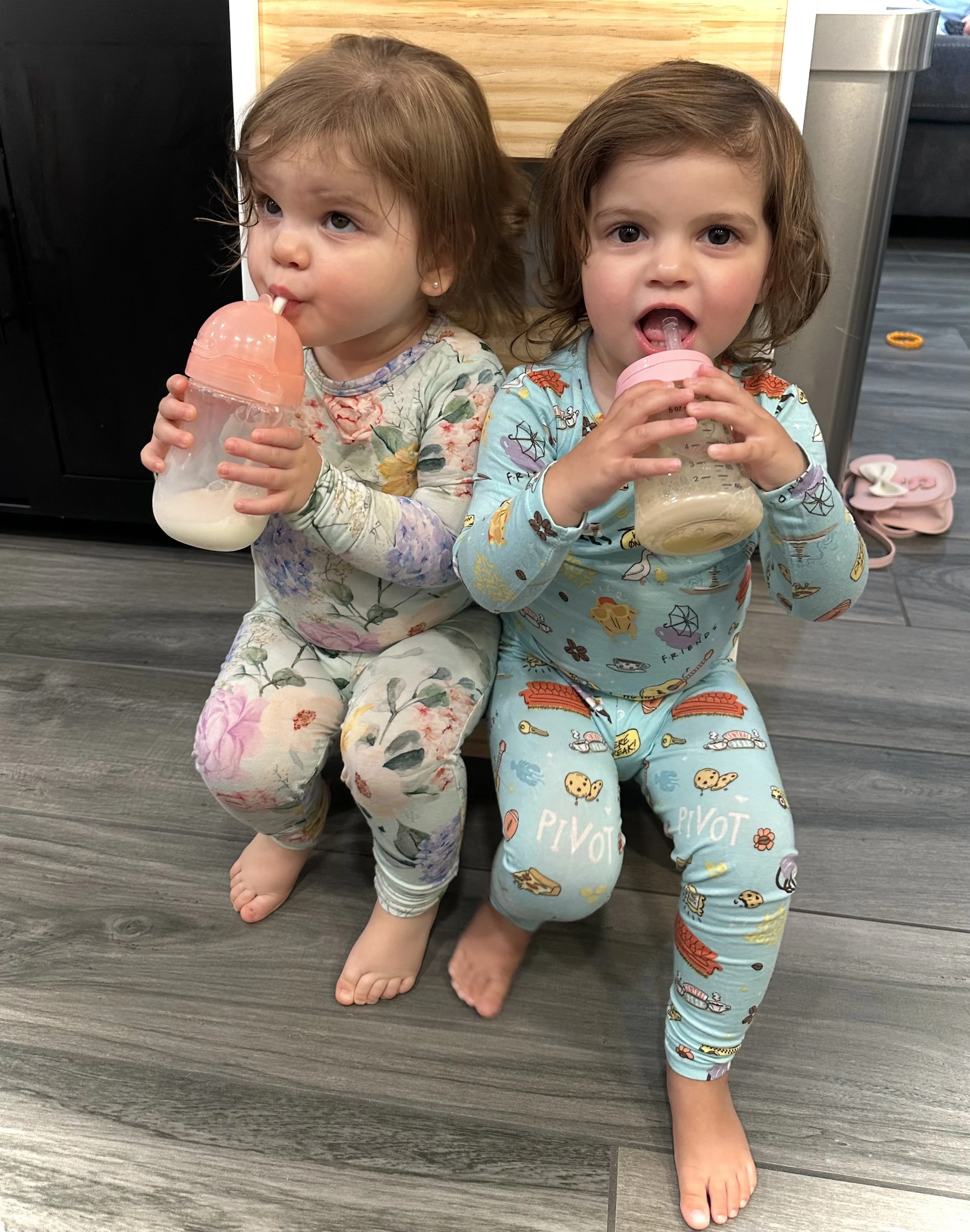 Two young girls sitting on the floor, each holding a sippy cup and drinking from it, in pajamas with colorful patterns.