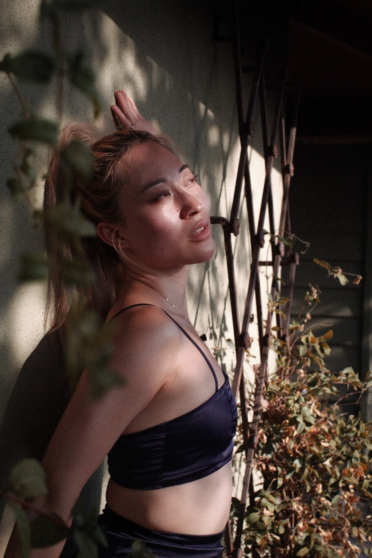 A woman with blonde hair and light skin lies on a wall, with her eyes partly closed, wearing a dark blue satin camisole. She is surrounded by green and brown leafy plants.