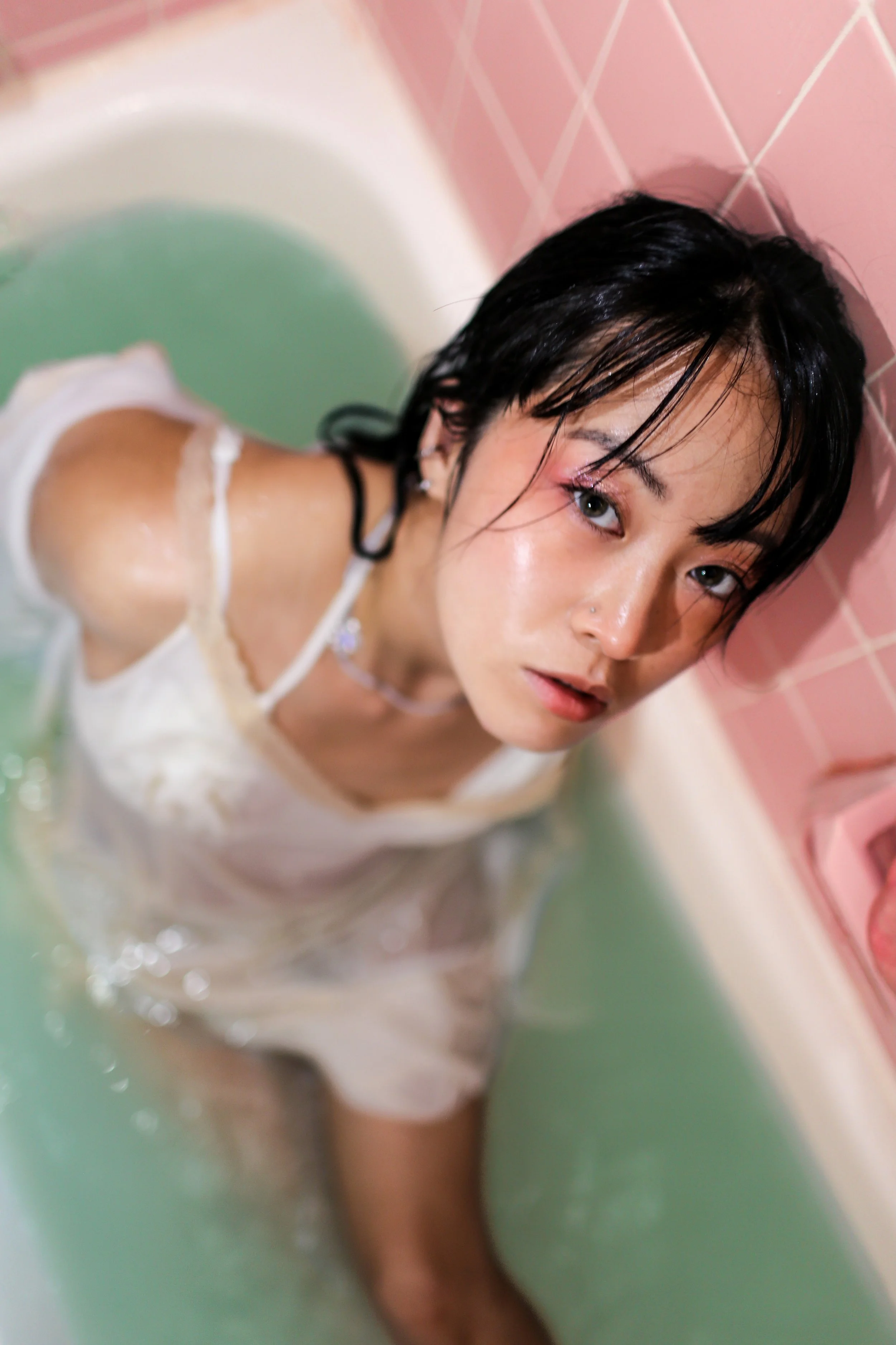 Woman with wet hair and makeup sitting in a bathtub filled with water, with pink tiled wall behind her.