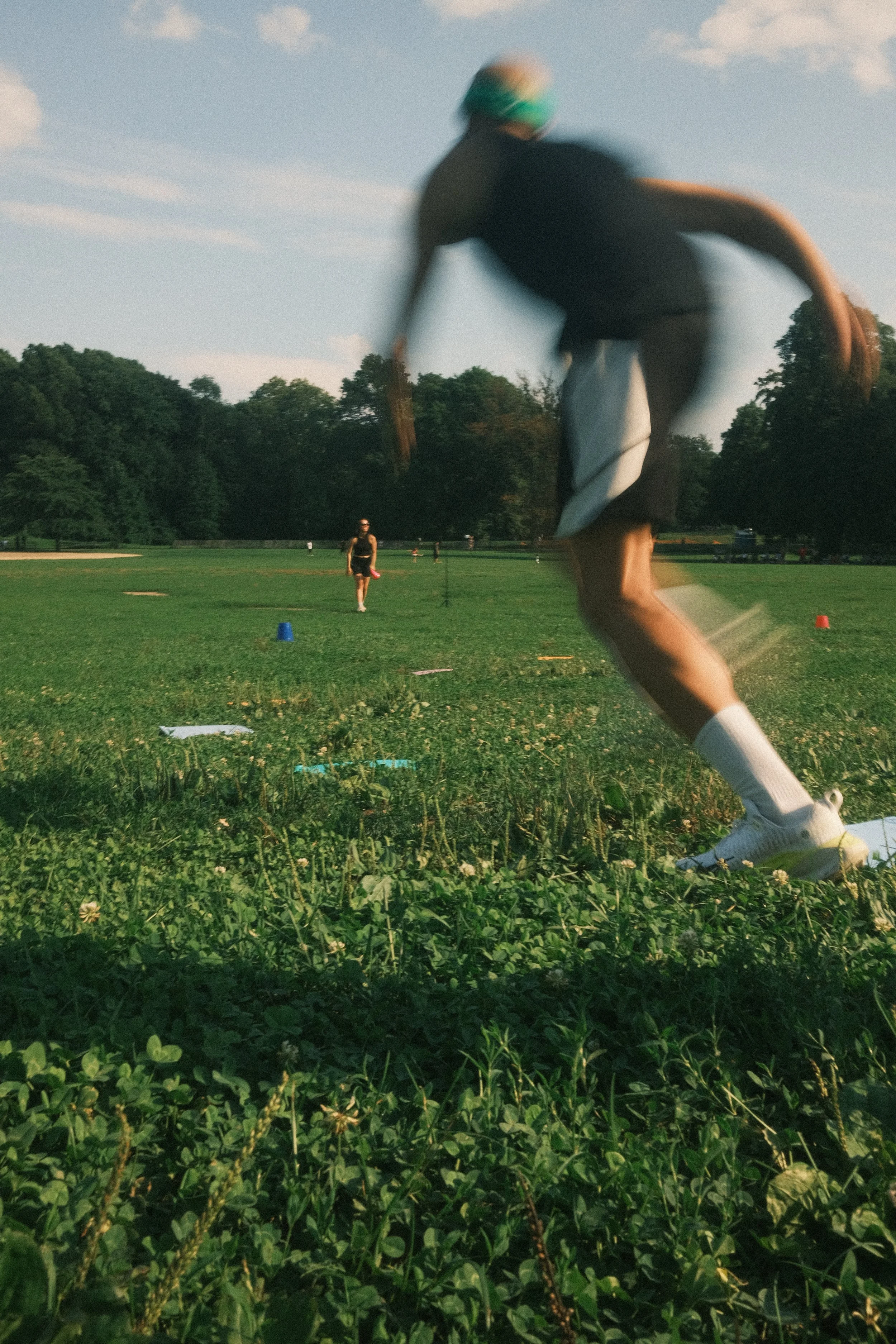 A person in athletic gear, wearing a swim cap and goggles, is running on a grassy field during daytime, with trees and a clear sky in the background.