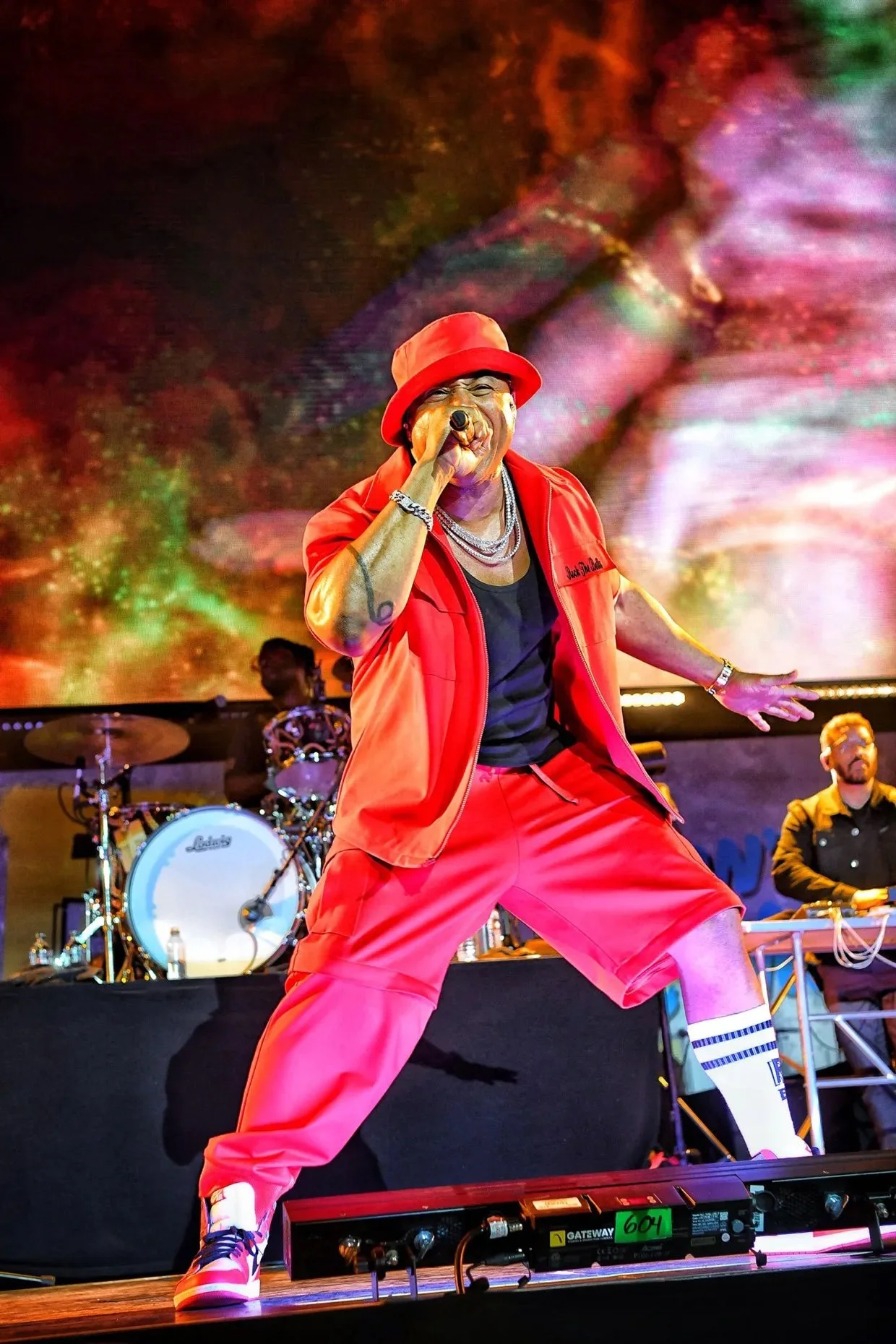 LL Cool J at Rock the Bells 2024 performing in a red outfit and hat rapping into a microphone on stage, with a colorful digital background and band members playing instruments behind him.