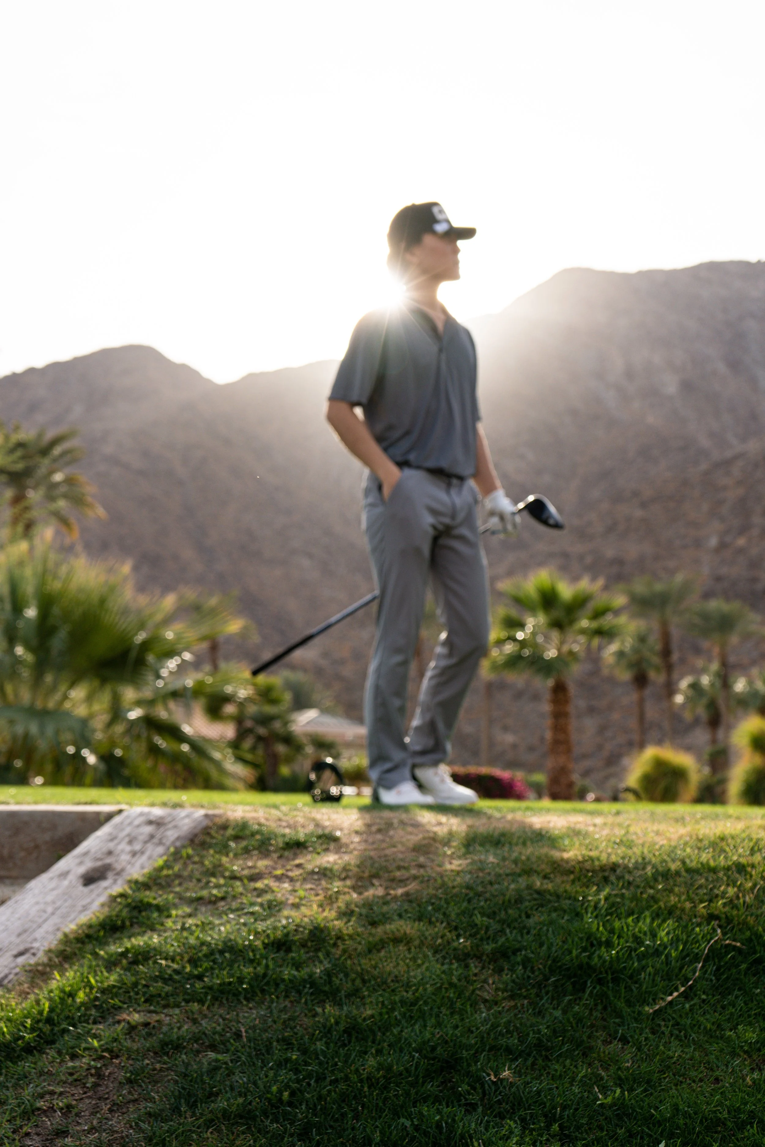A person standing on a green golf course in Palm Desert with mountains in the background during sunset, holding a golf club and wearing casual golf attire.