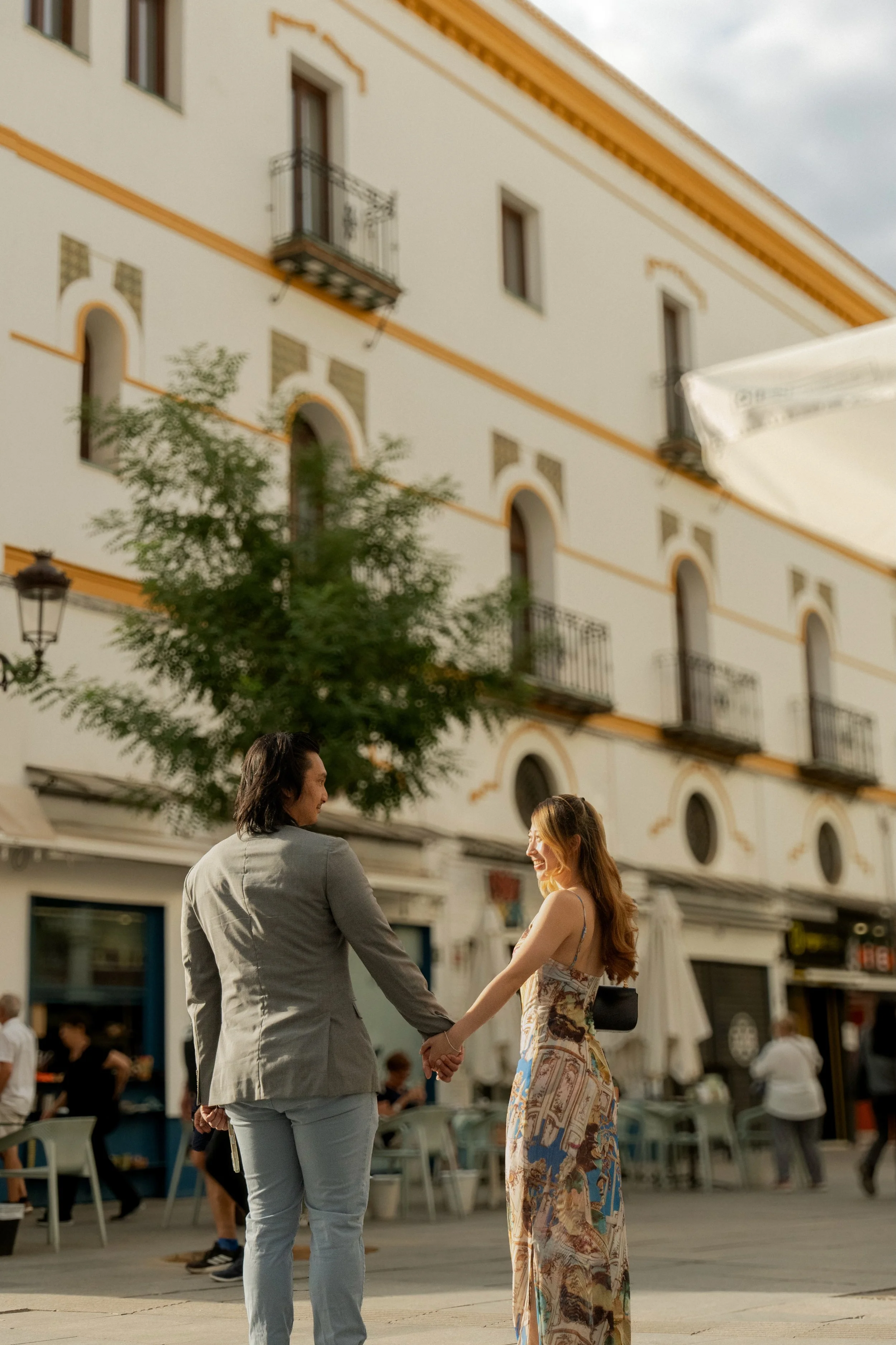 A man and woman holding hands and smiling at each other outdoors in front of a white building in Seville, Spain, with yellow trim, balconies, and arched windows, with other people in the background.