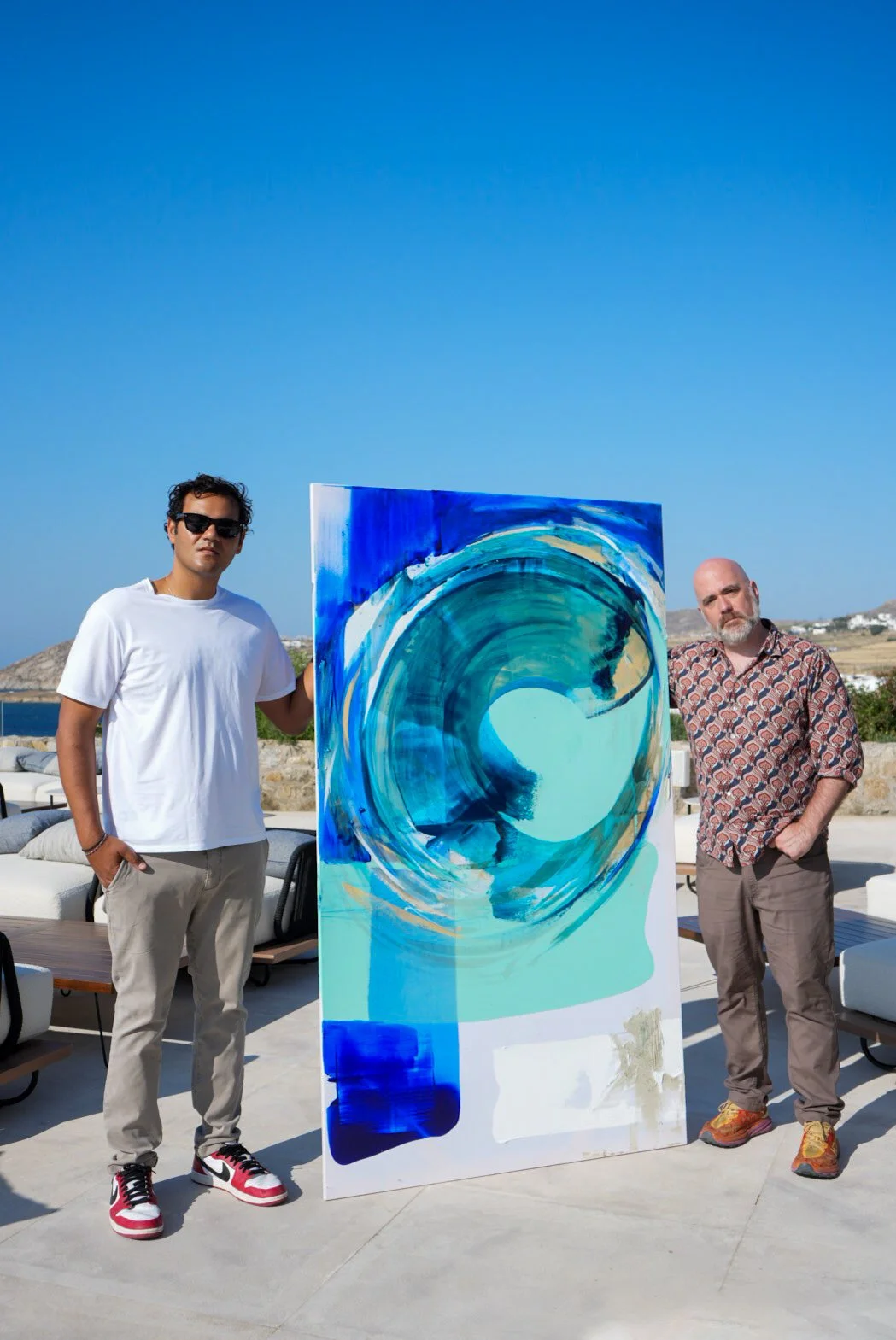 Two men stand on a patio with a large abstract painting of a blue wave in front of them. One man is wearing a white t-shirt and sunglasses, the other a patterned shirt and tan pants. The background shows outdoor furniture, a hillside, and a clear blue sky.