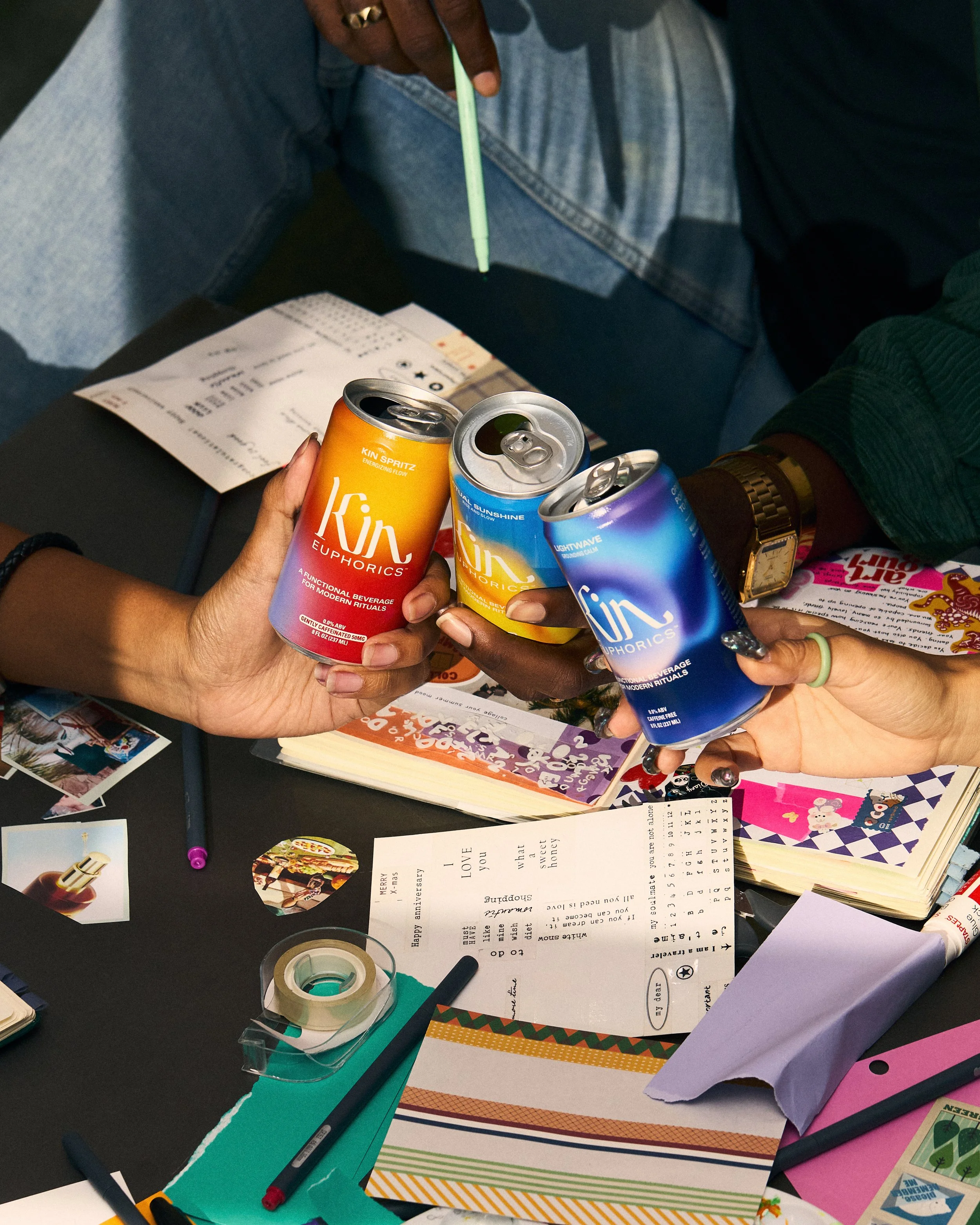 Multiple hands holding colorful drink cans titled Kins on a cluttered desk with papers, pens, and stationery.
