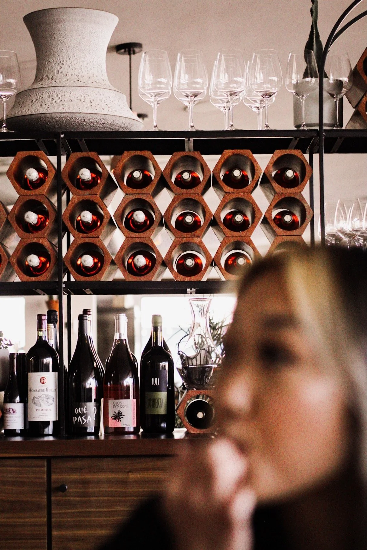 A bar shelf holding wine bottles, glasses, and a large white decorative vase. The wine bottles are stored in a honeycomb wine rack with a mix of red wines and informal labels. Several empty wine glasses are placed on the upper shelf, and some bottles