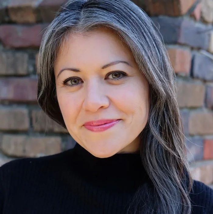 Head and shoulder shot of a middle aged woman with fair skin, green eyes, and brown hair with grey streaks, wearing a black turtleneck sweater in front of a brick background