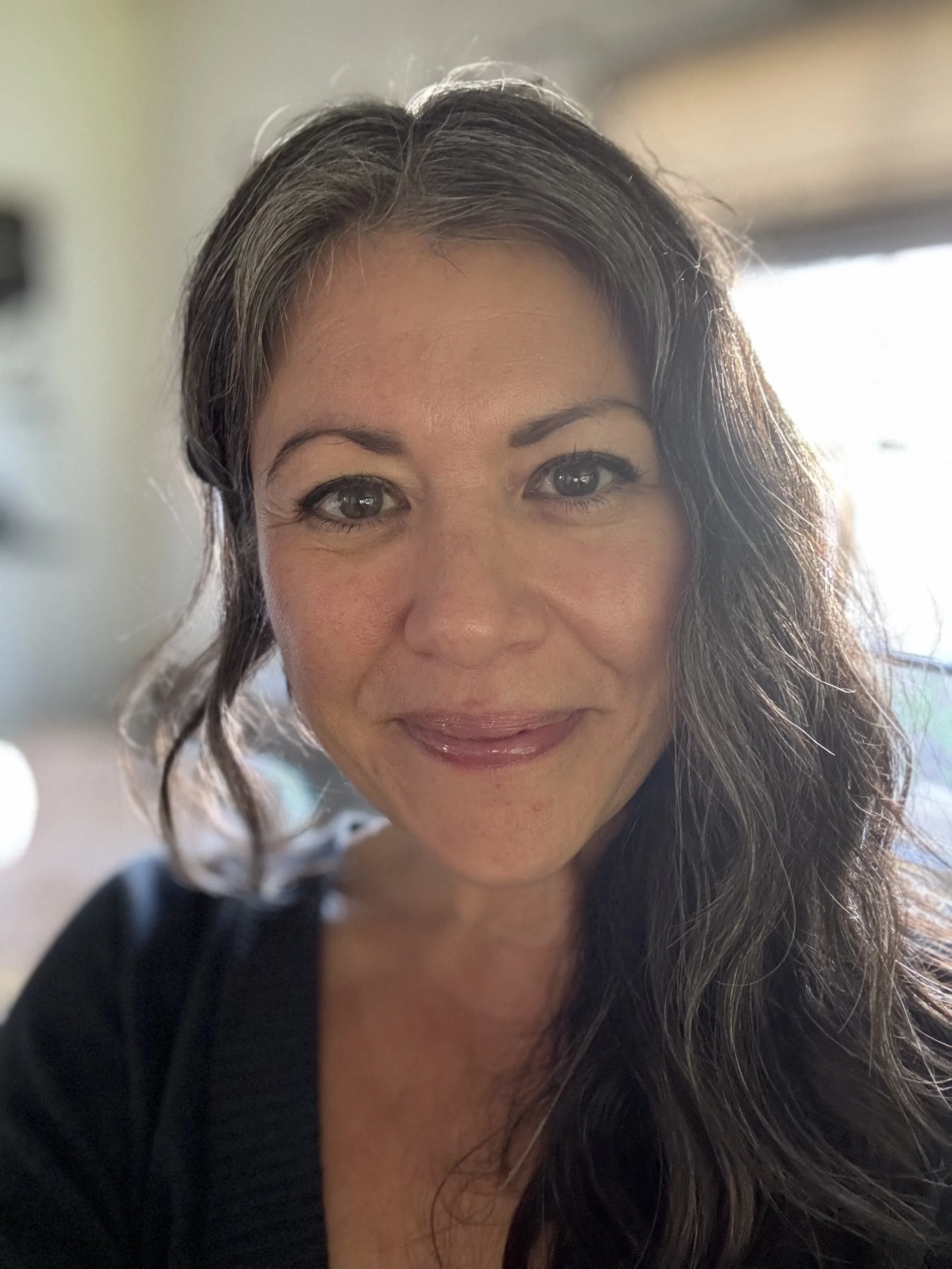 Woman with green eyes, long, wavy, dark brown and grey hair, wearing a v-neck blouse, smiling with mouth closed and blurred out window in the background