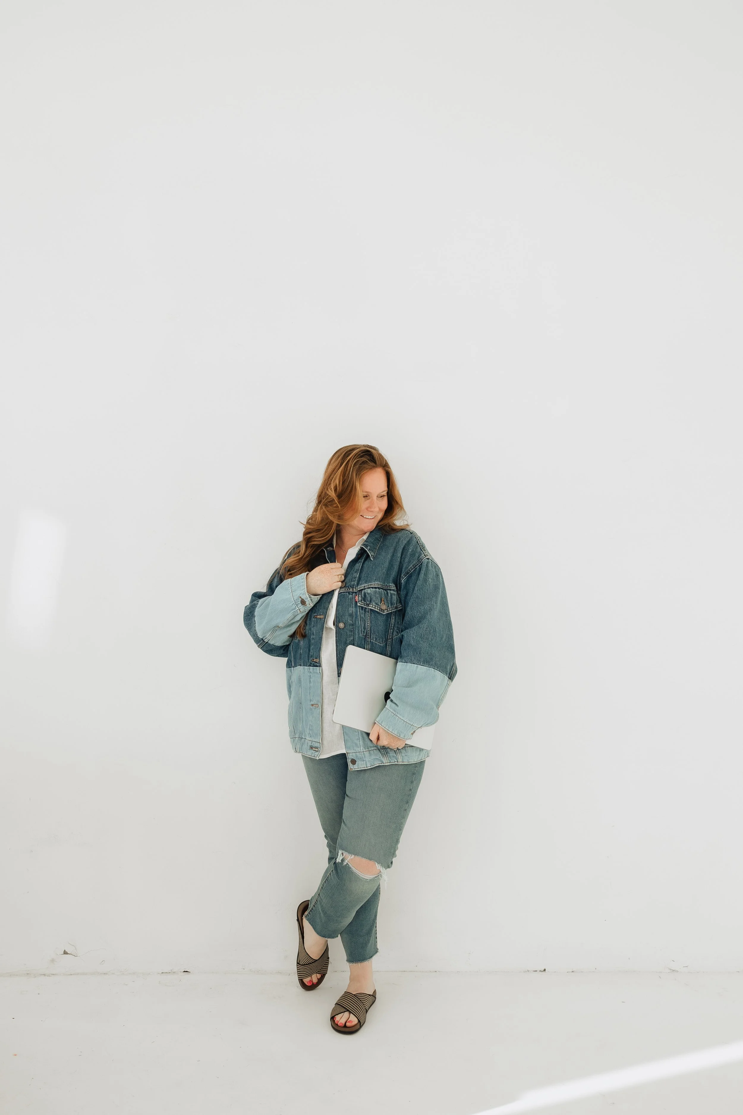 A woman with long brown hair wearing a denim jacket, jeans with a ripped knee, and sandals, holding a closed laptop, standing against a plain white wall.