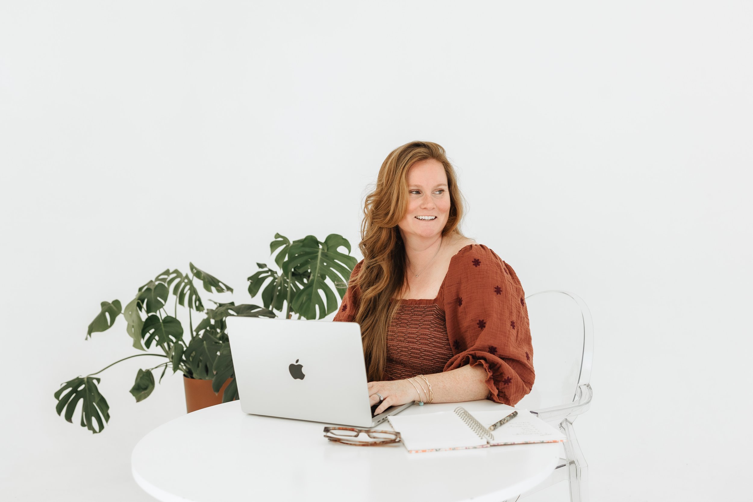 A woman with long red hair sitting at a white desk, using a silver MacBook laptop, with a large green potted plant behind her, dressed in a rust-colored blouse with puffed sleeves.