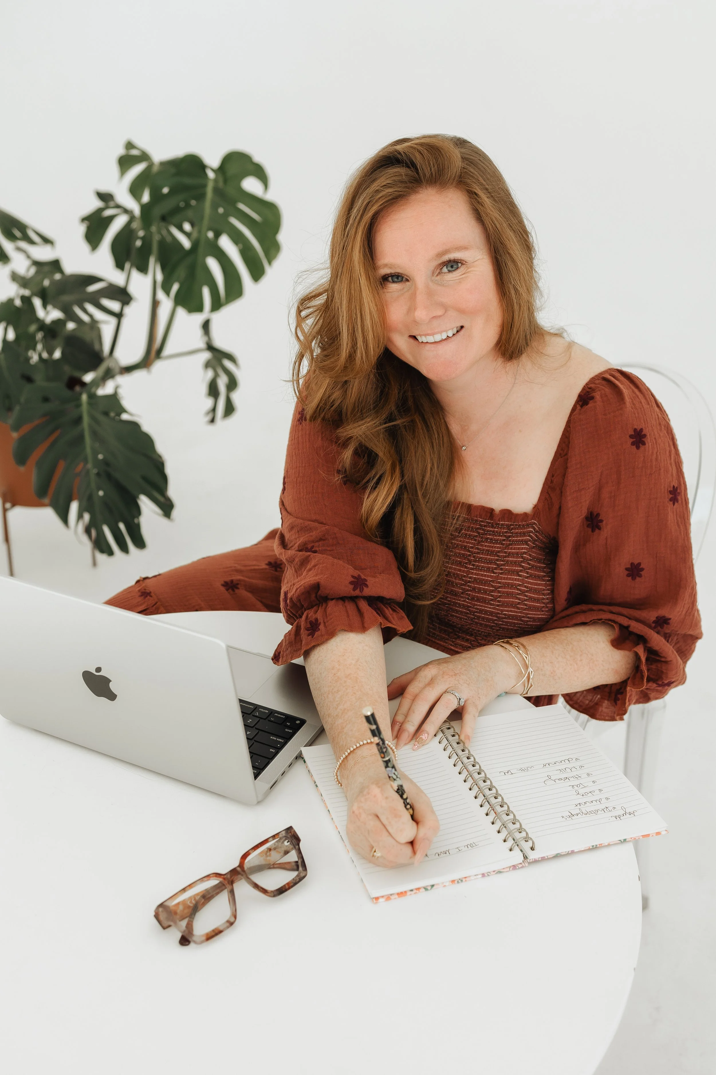 A woman with red hair, wearing a rust-colored dress with floral embroidery, sits at a white table with a MacBook, a spiral notebook, and a pair of glasses, smiling and writing in her notebook, with a large green houseplant in the background.