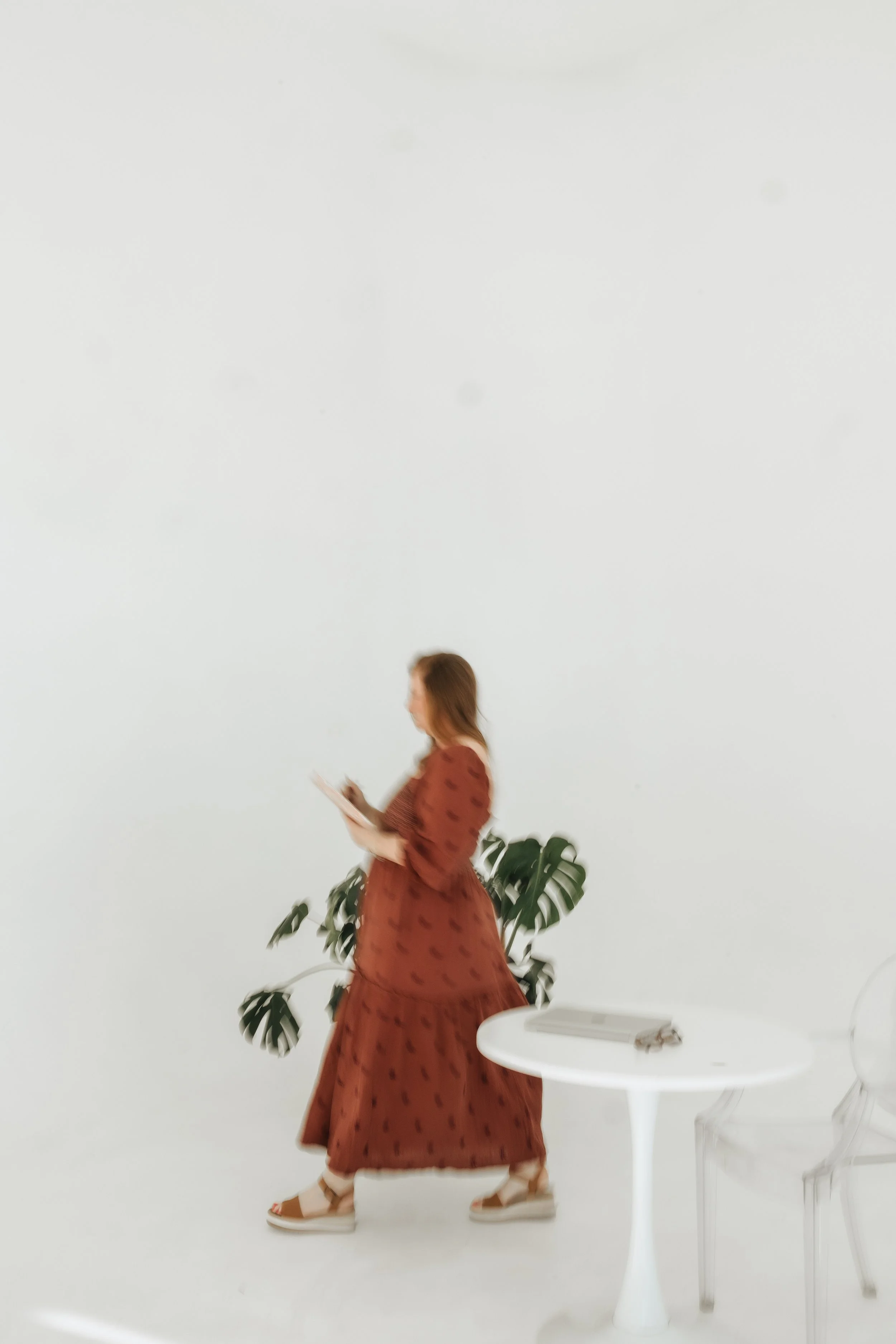 A woman in a long, brown dress with a pattern, holding a notebook and pen, standing near a leafy green plant, with a white table and clear chairs in a minimalistic white space.