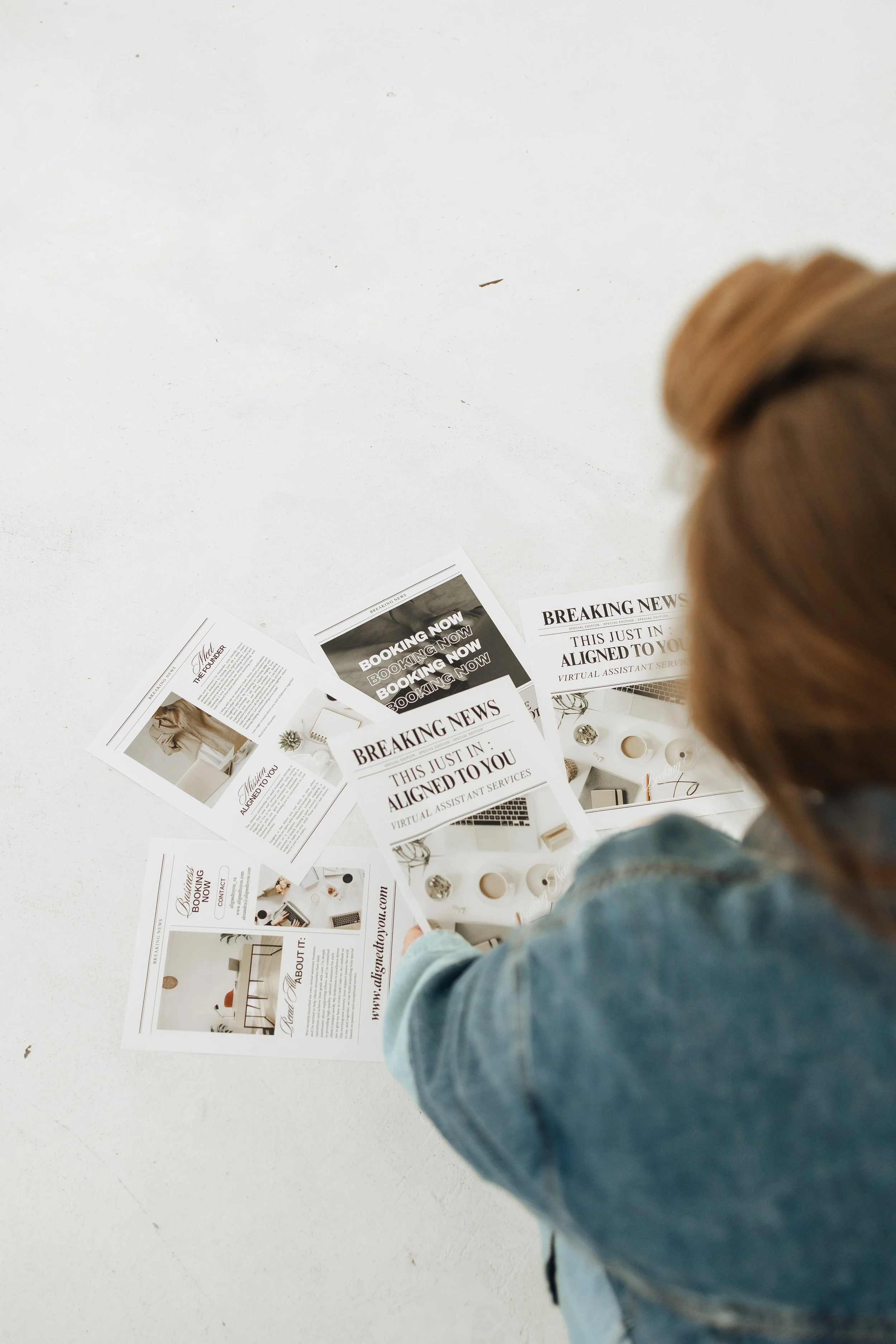 Person with reddish hair in a bun wearing a denim jacket looking at various printed flyers and brochures on a white surface.