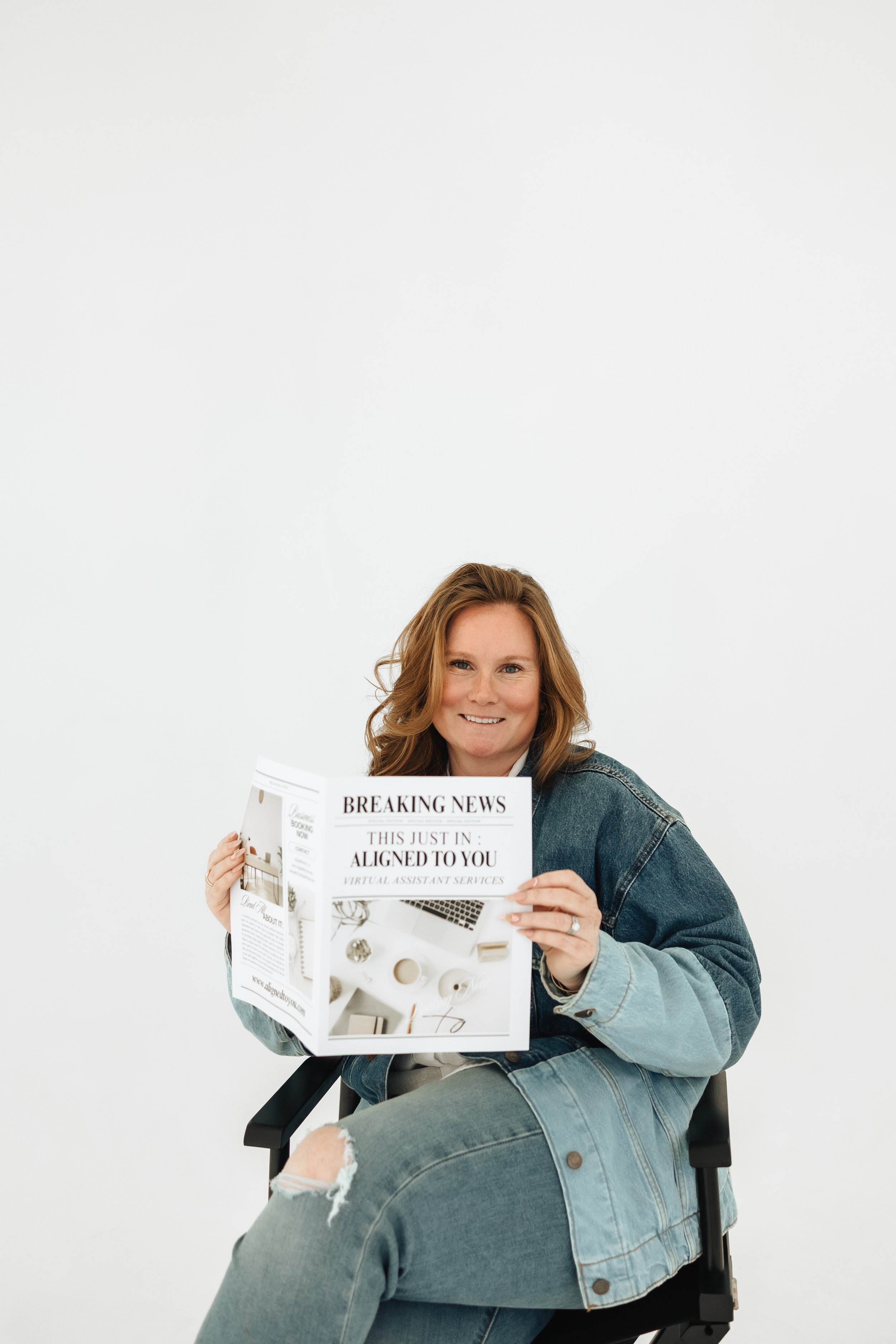 Woman sitting in a chair holding a newspaper with the headline 'Breaking News' and smiling at the camera, against a plain white background.