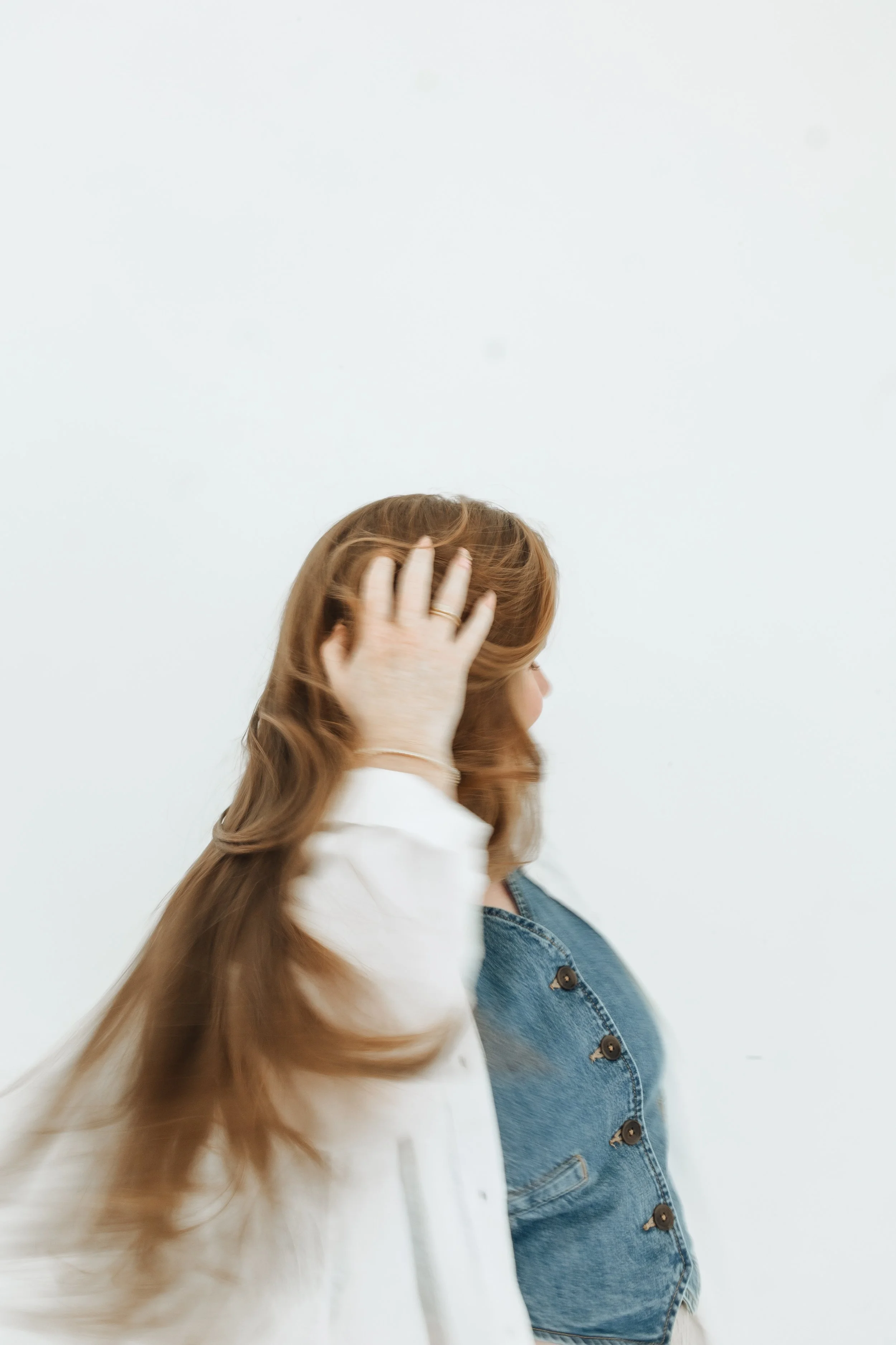 A woman with long, wavy, reddish-brown hair, wearing a denim jacket, faces to the right with her head slightly turn away, touching her hair with her left hand, against a plain white background.