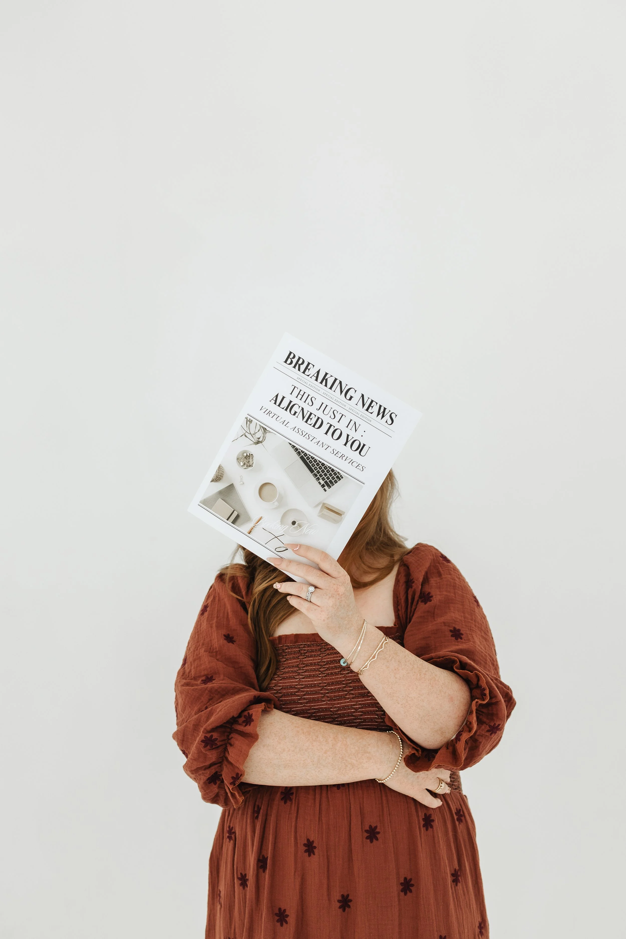 A woman in a rust-colored dress with black embroidered flowers is holding a magazine with a newspaper headline in front of her face. The background is plain white.