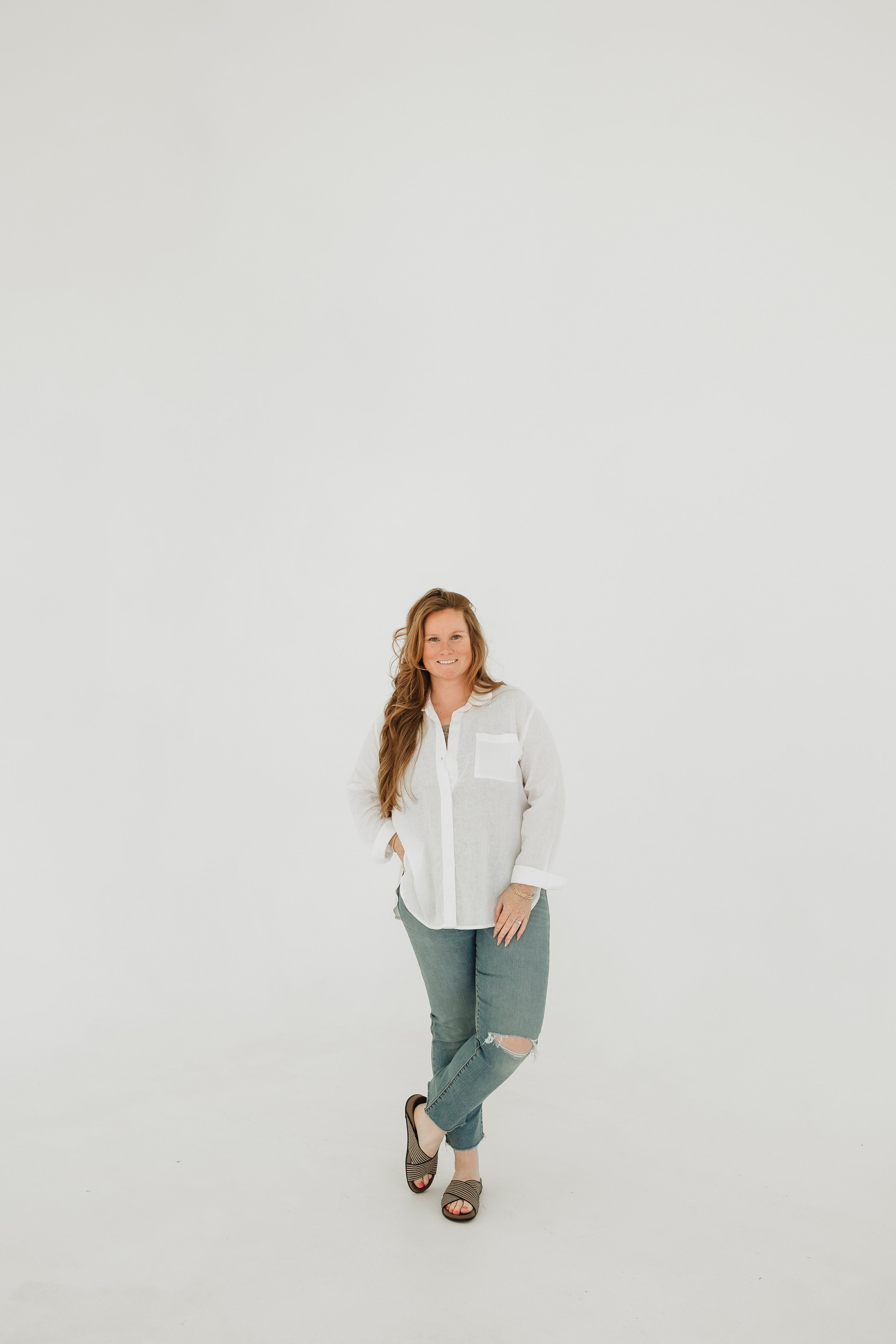 A woman standing in a studio with a white background, wearing a white shirt, ripped jeans, and open-toe shoes, smiling and looking at the camera.