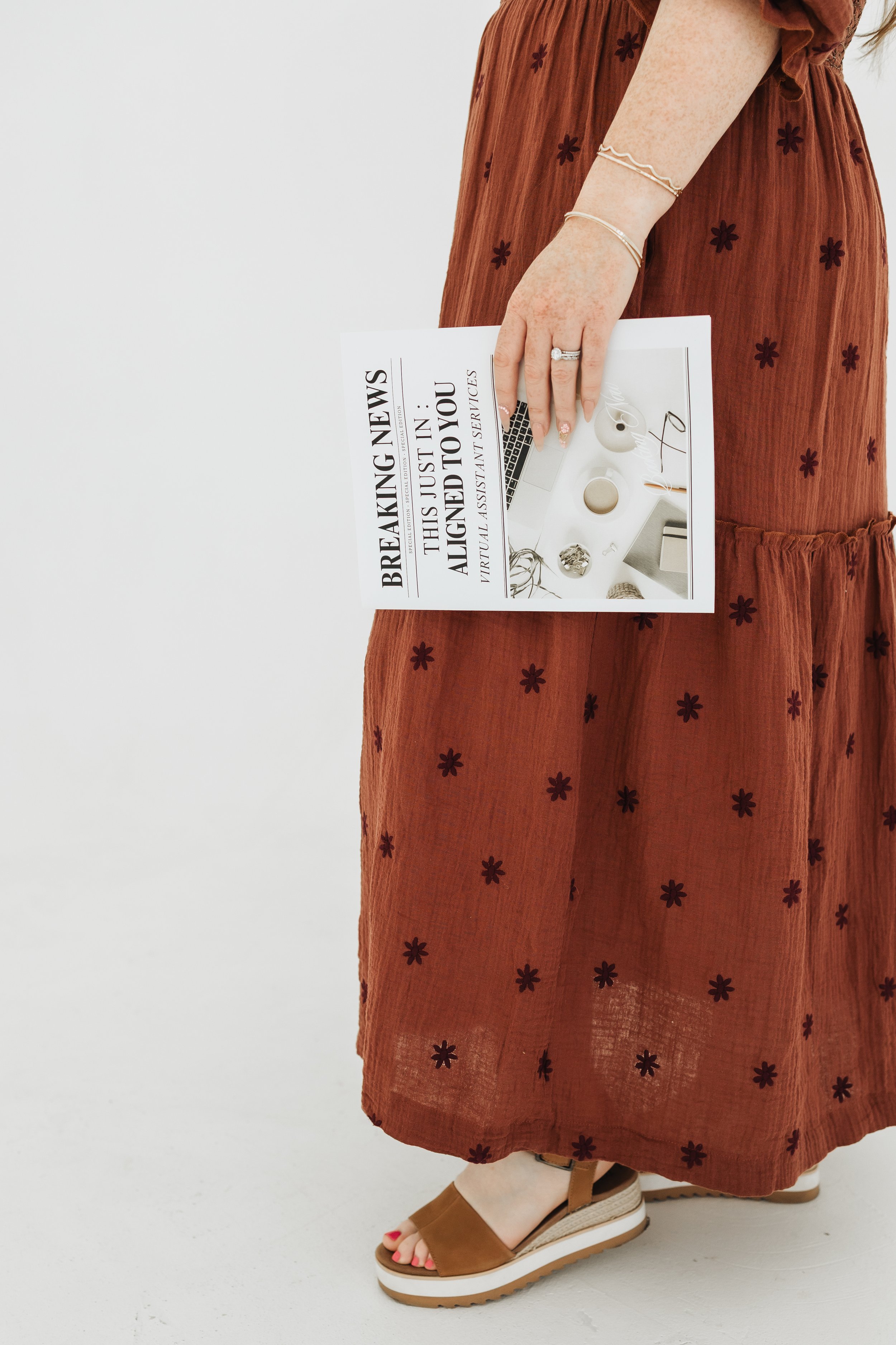 A woman in a rust-colored dress with black flower patterns is holding a newspaper or magazine. Part of her right arm and hand are visible, showing jewelry including a ring and bracelets. She is wearing sandals with wedge heels and has red nail polish.