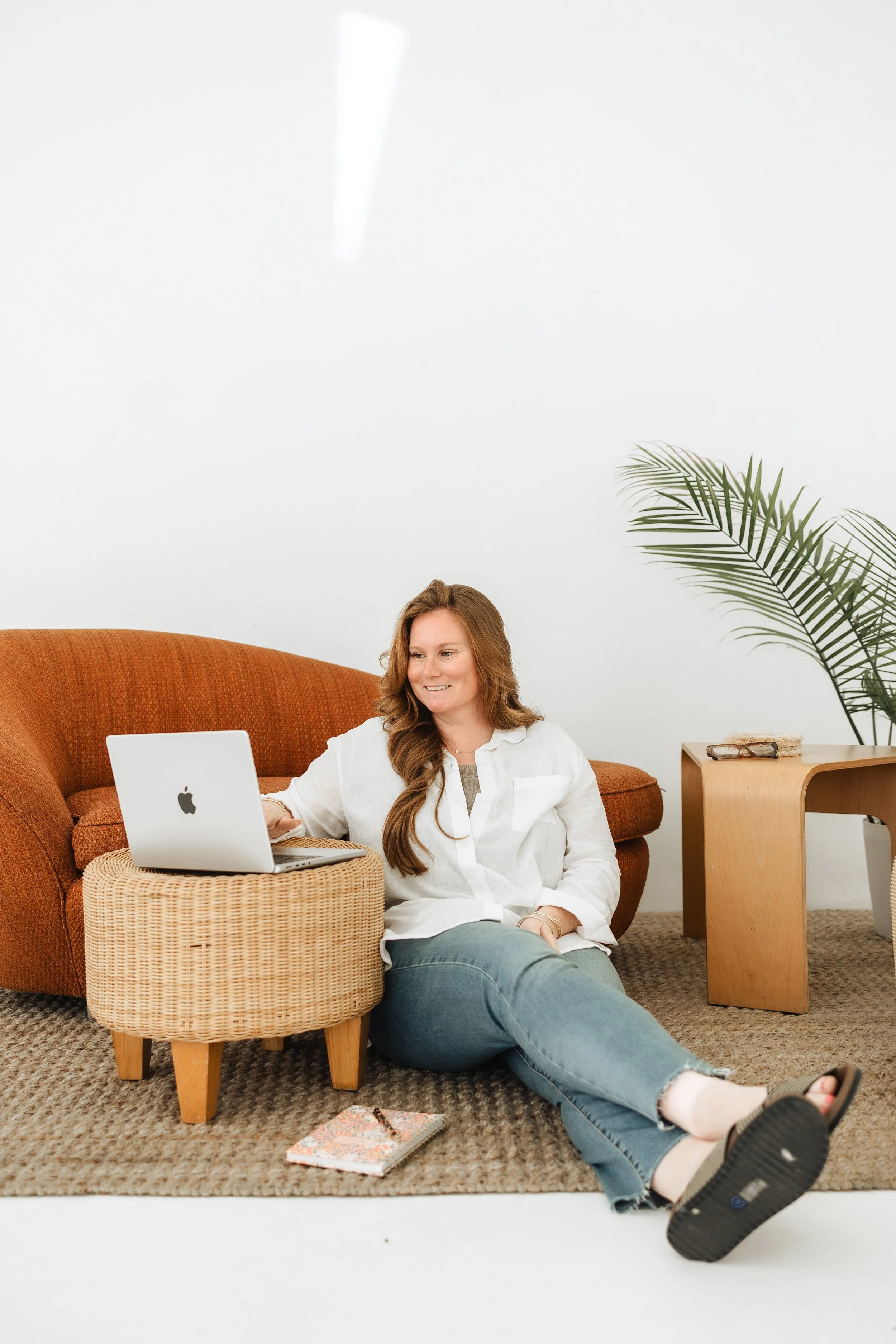 Woman sitting on the floor with a laptop on a wicker stool, in a minimalist room with a white wall, a orange couch, a wooden side table, a plant, and a notebook on the floor.