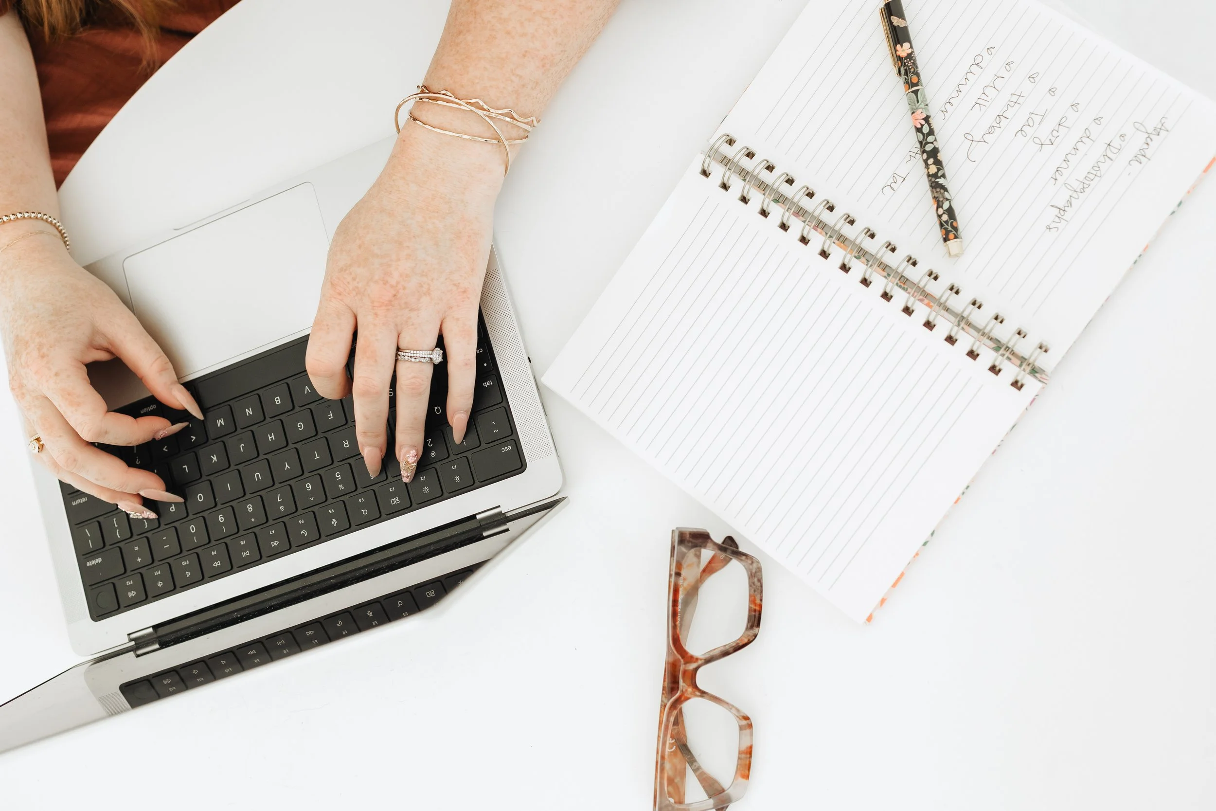 Person typing on a laptop with a notebook, a pen, and a pair of glasses on a white desk.