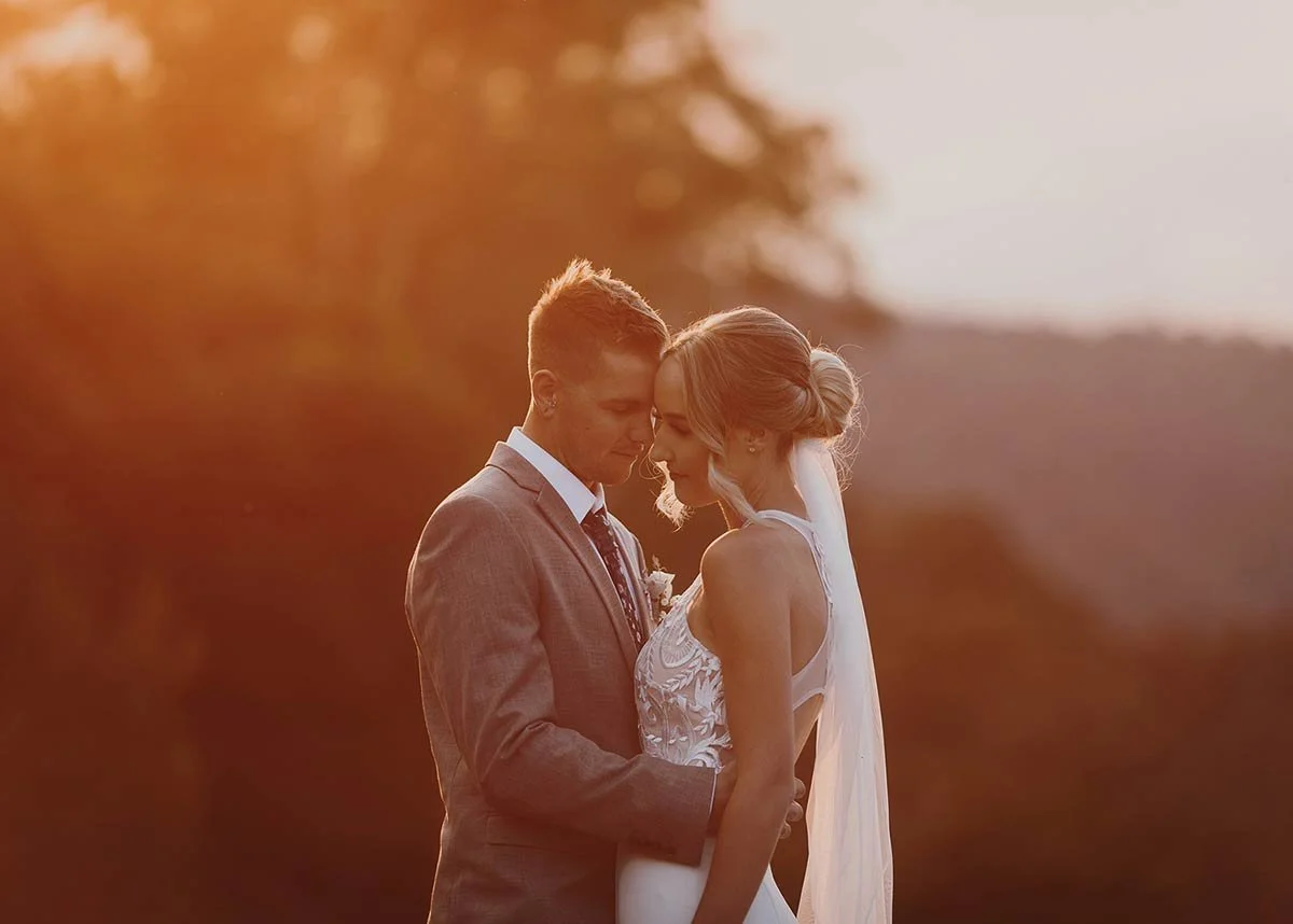 A bride and groom share a tender moment outdoors during sunset, with their foreheads touching and eyes closed.