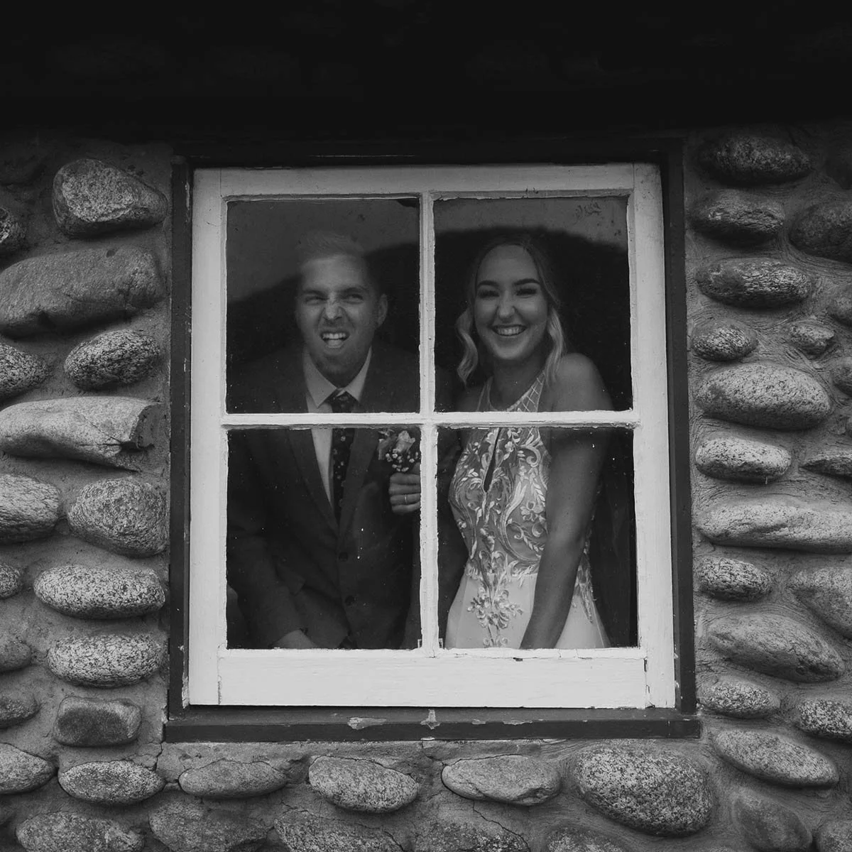A couple smiling and making silly faces behind a window with four panes, with a stone wall exterior.