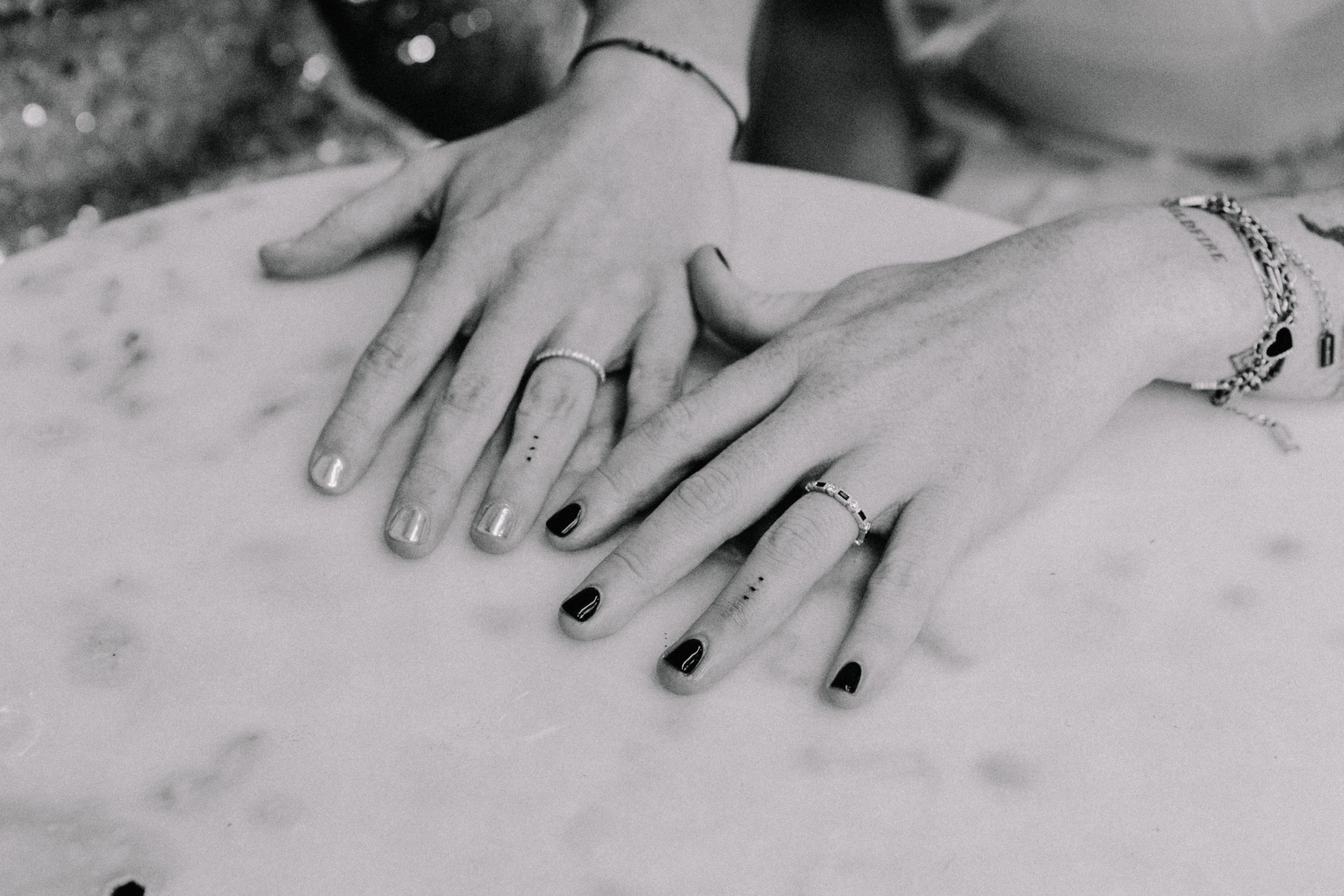 Two hands with rings, bracelets, and painted nails are placed on a surface, overlapping each other in a close-up shot in black and white.