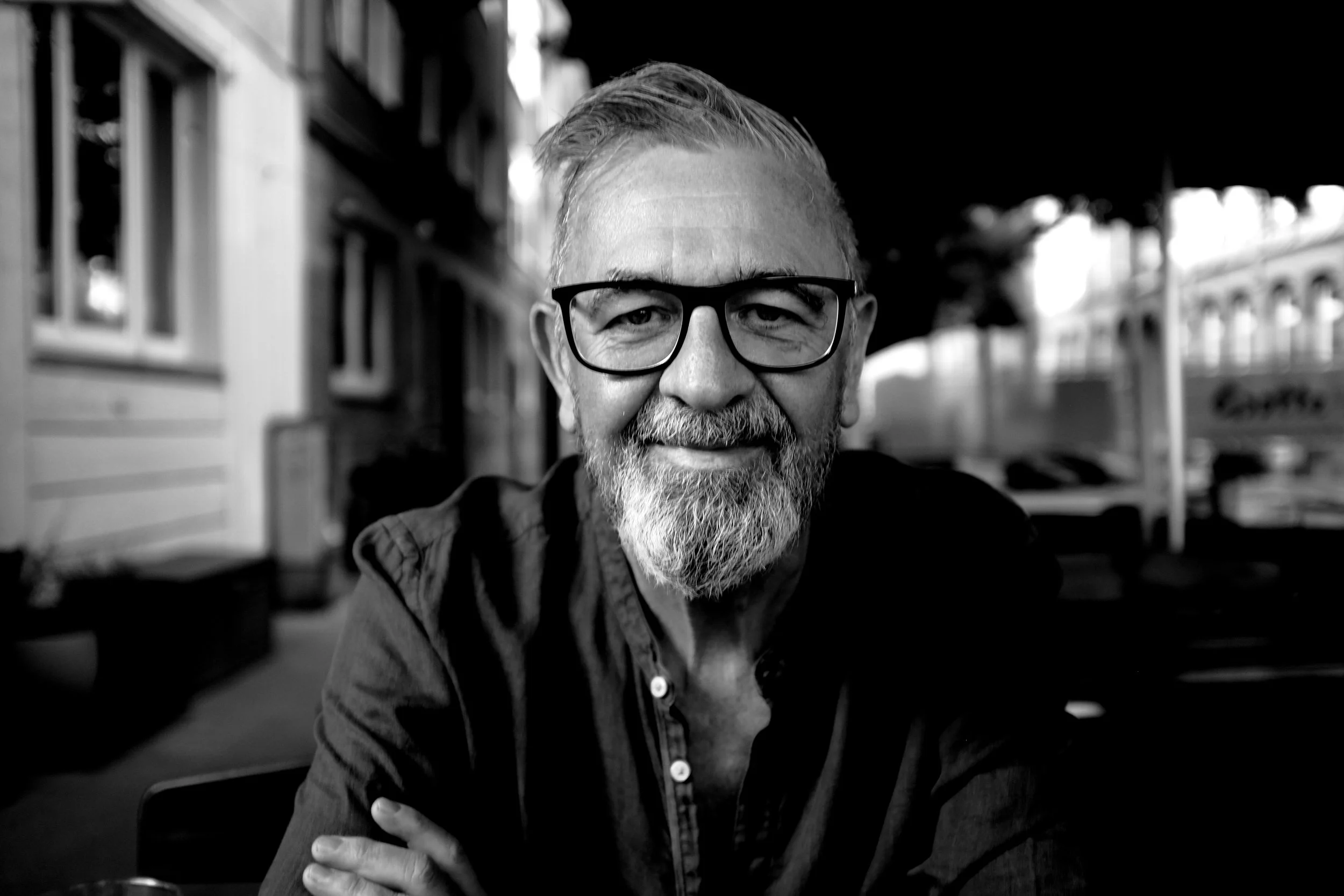 Schwarz-weiß-Foto eines lächelnden Mannes mit Brille, grauem Bart und kurzen Haaren, sitzend im Freien.