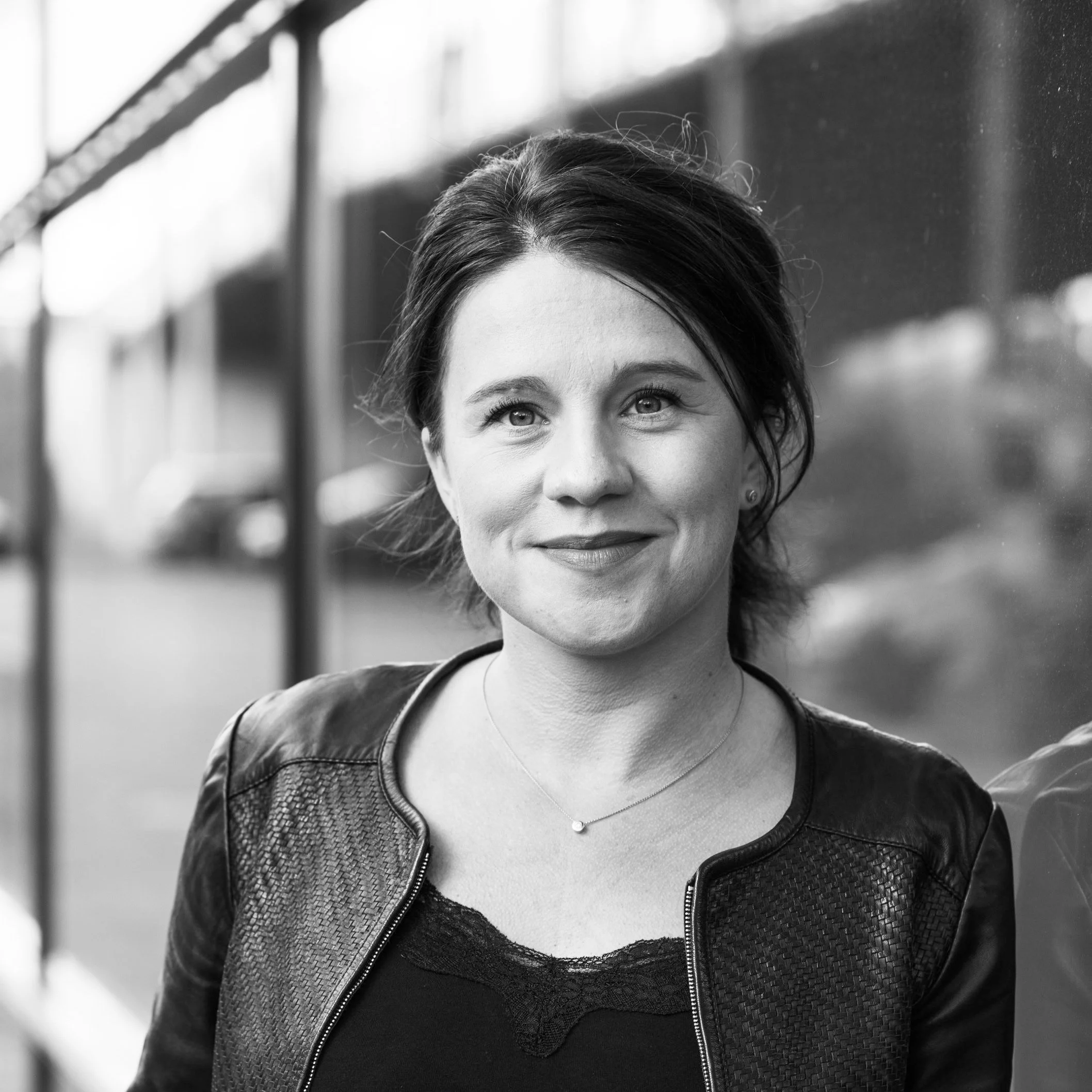 Schwarz-weiß-Fotografie einer lächelnden Frau mit dunklem Haar, Lederjacke und Schmuck vor einer Glaswand