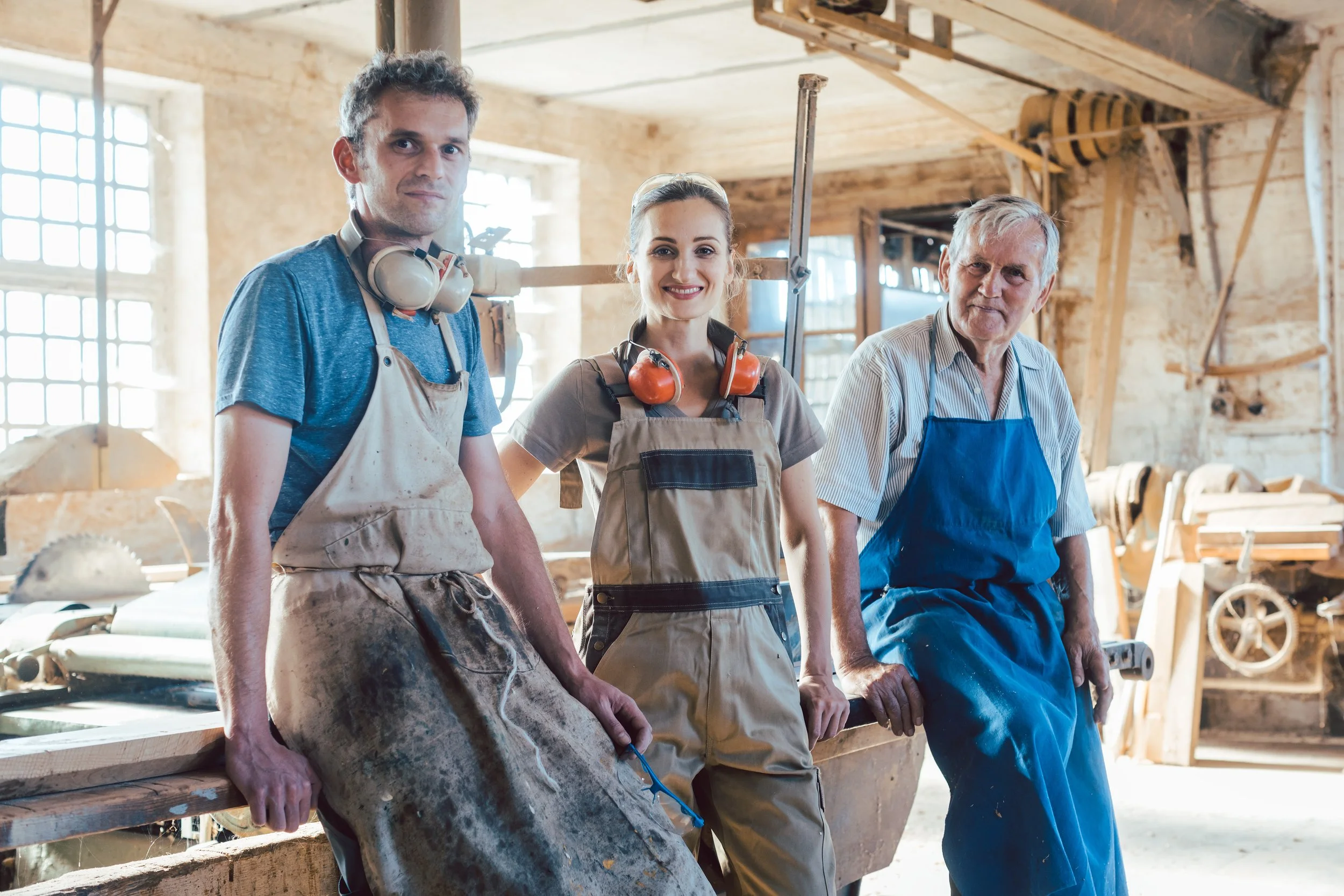 Drei Menschen in einer Werkstatt, die Werkzeuge und Baumaterialien nutzen, tragen Schutzkleidung und stehen vor einem Arbeitsbereich mit Holzbrettern und Werkzeugen.