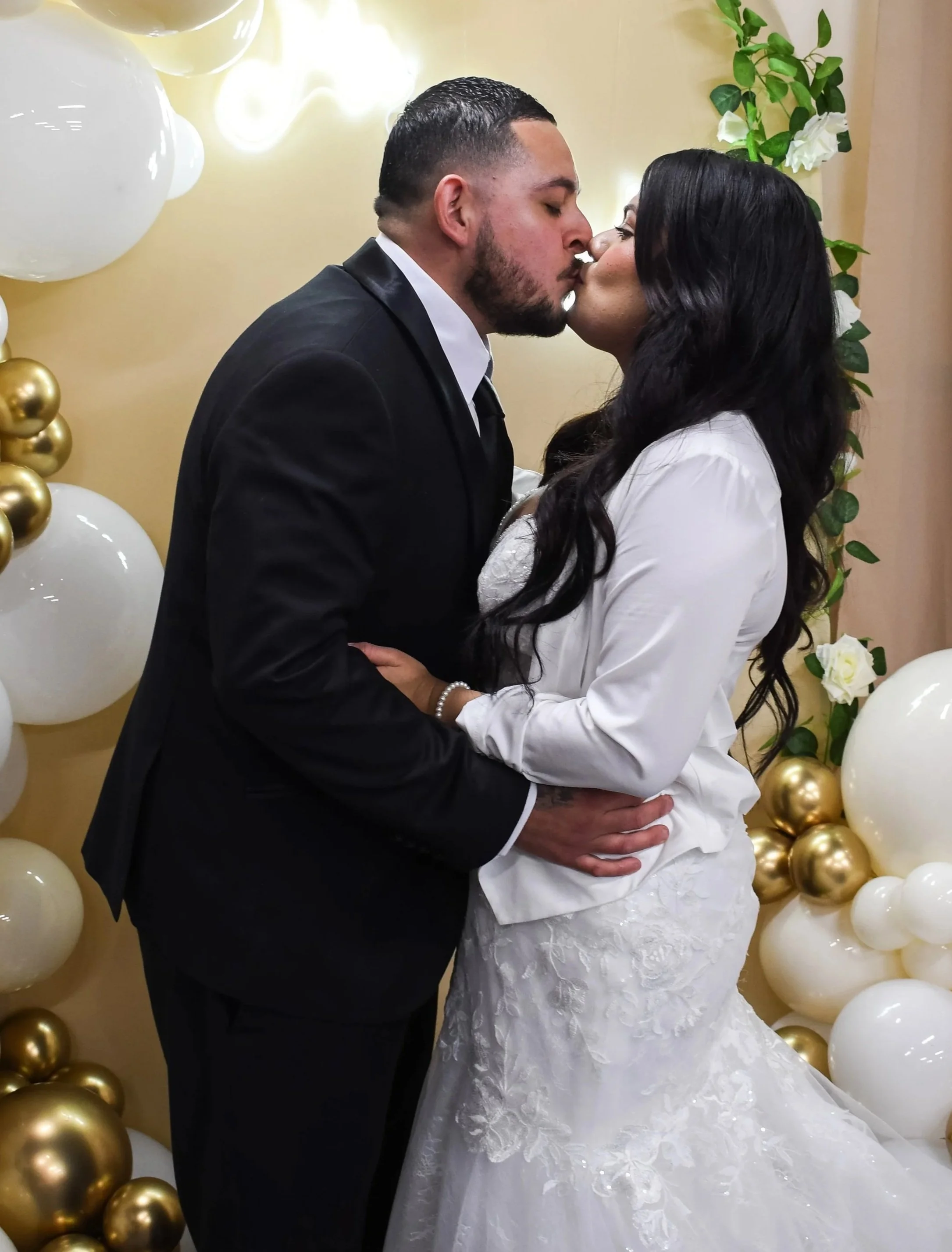 A couple kissing at their wedding reception, standing in front of gold and white balloons and greenery decoration.