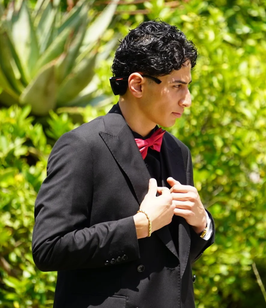 A young man dressed in a black suit with a red bow tie, wearing glasses, standing outdoors amid lush green plants, adjusting his suit.