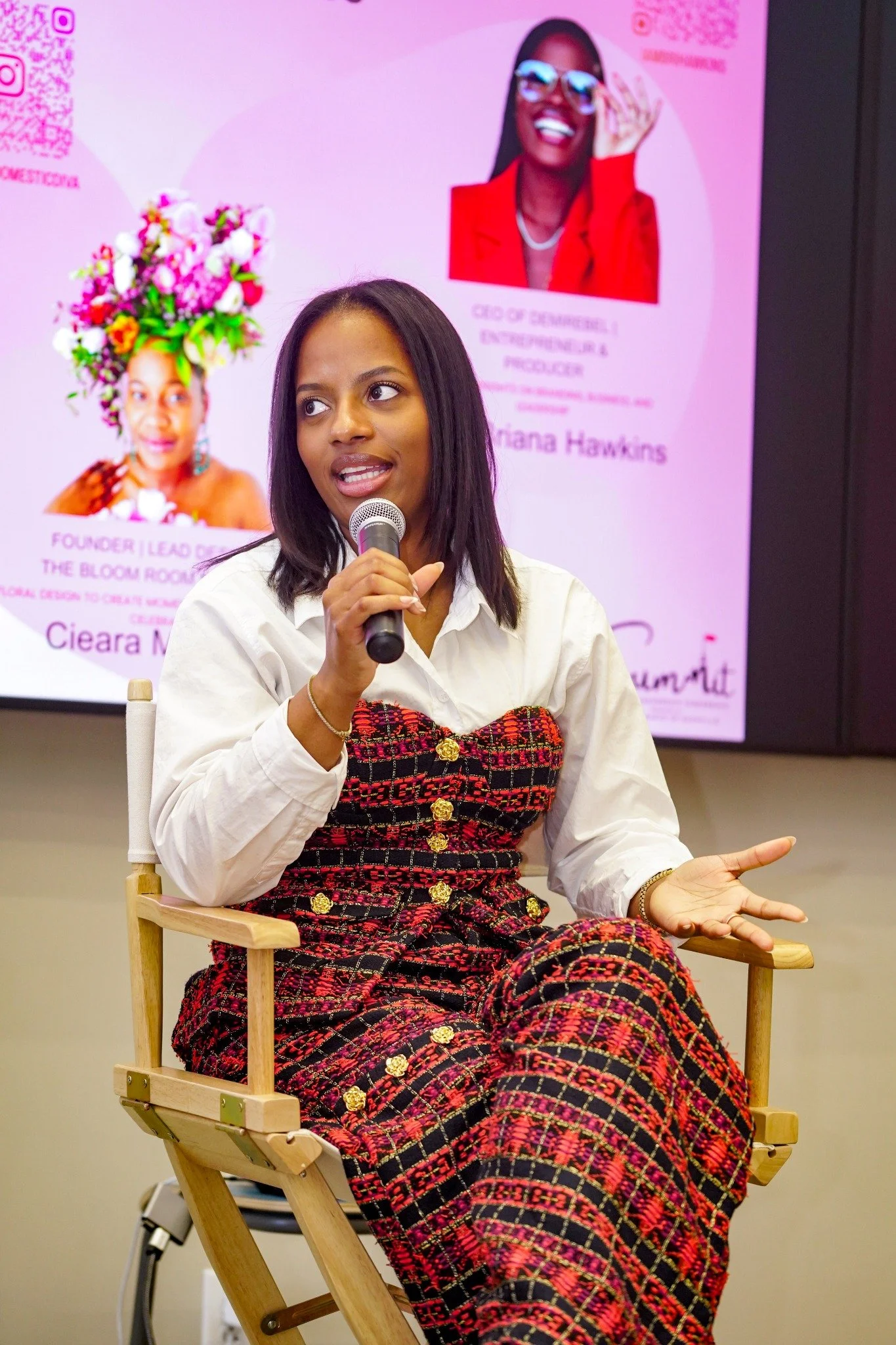 Briana Hawkins speaking into a microphone during a panel conversation, wearing a red and black patterned outfit.