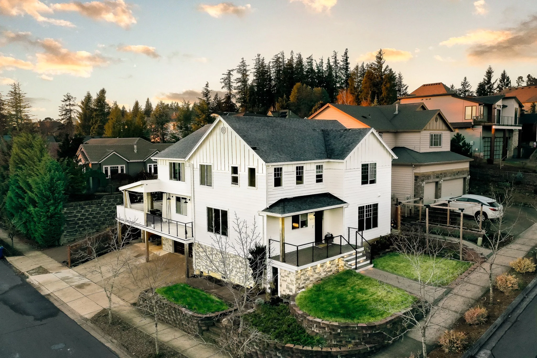 A white two-story house with a dark roof, multiple windows, and a small balcony in a neighborhood. The house has a stone foundation, a small front lawn, and a paved sidewalk, with neighboring houses and trees around.
