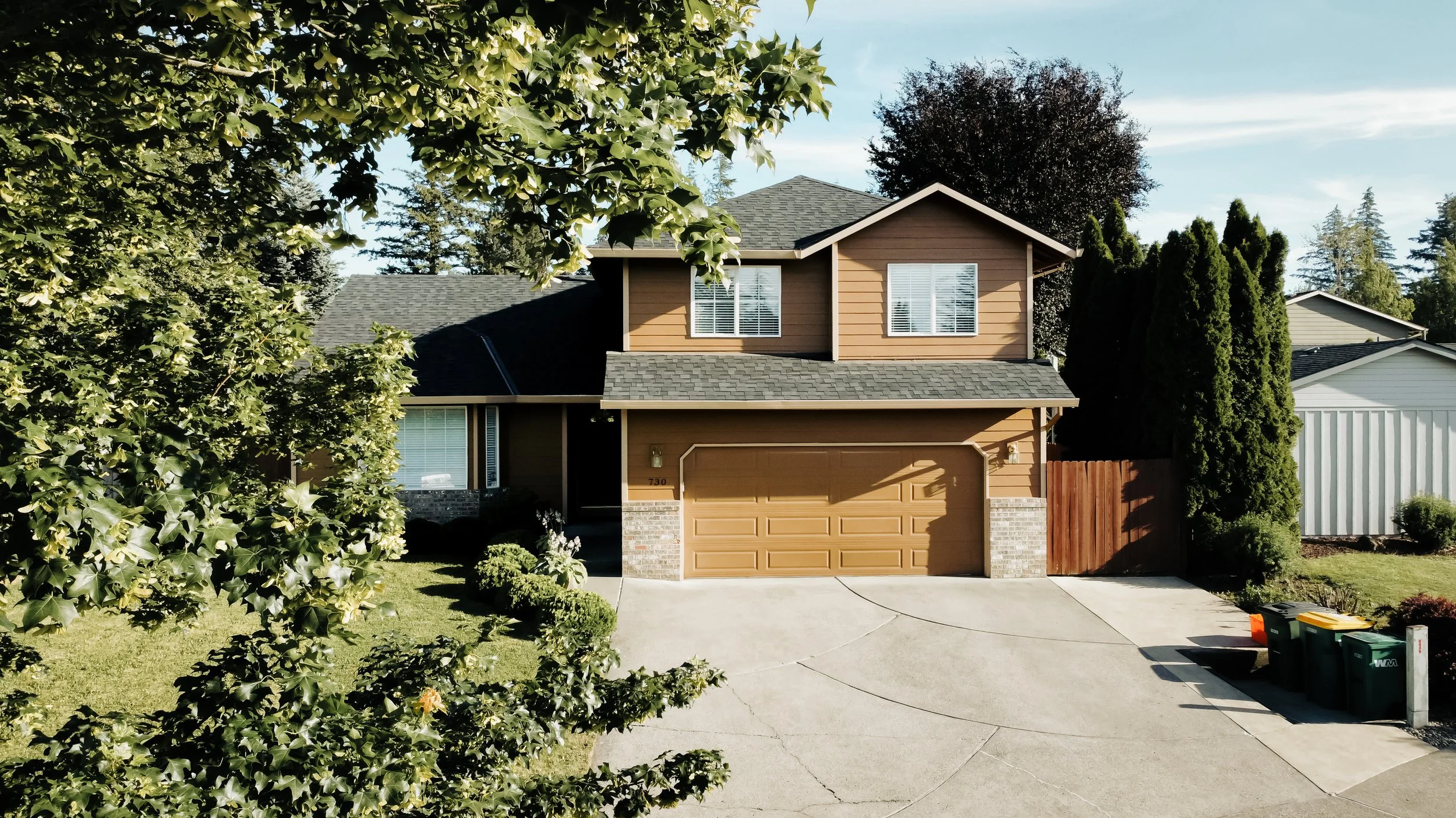 A two-story house with a tan wooden exterior, black roof, and attached garage, surrounded by lush green trees and a well-maintained lawn, with some trash bins on the driveway.