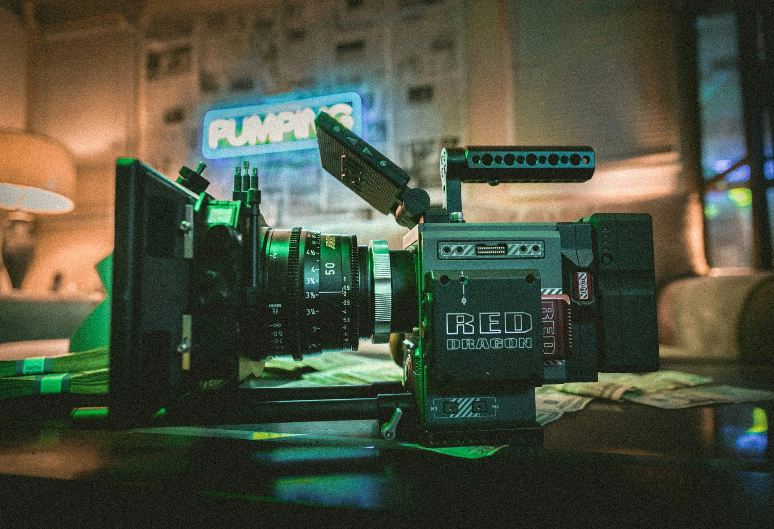Red Dragon cinema camera on a table with film money underneath, in a dimly lit room with a neon sign reading "PUMPING" in the background.